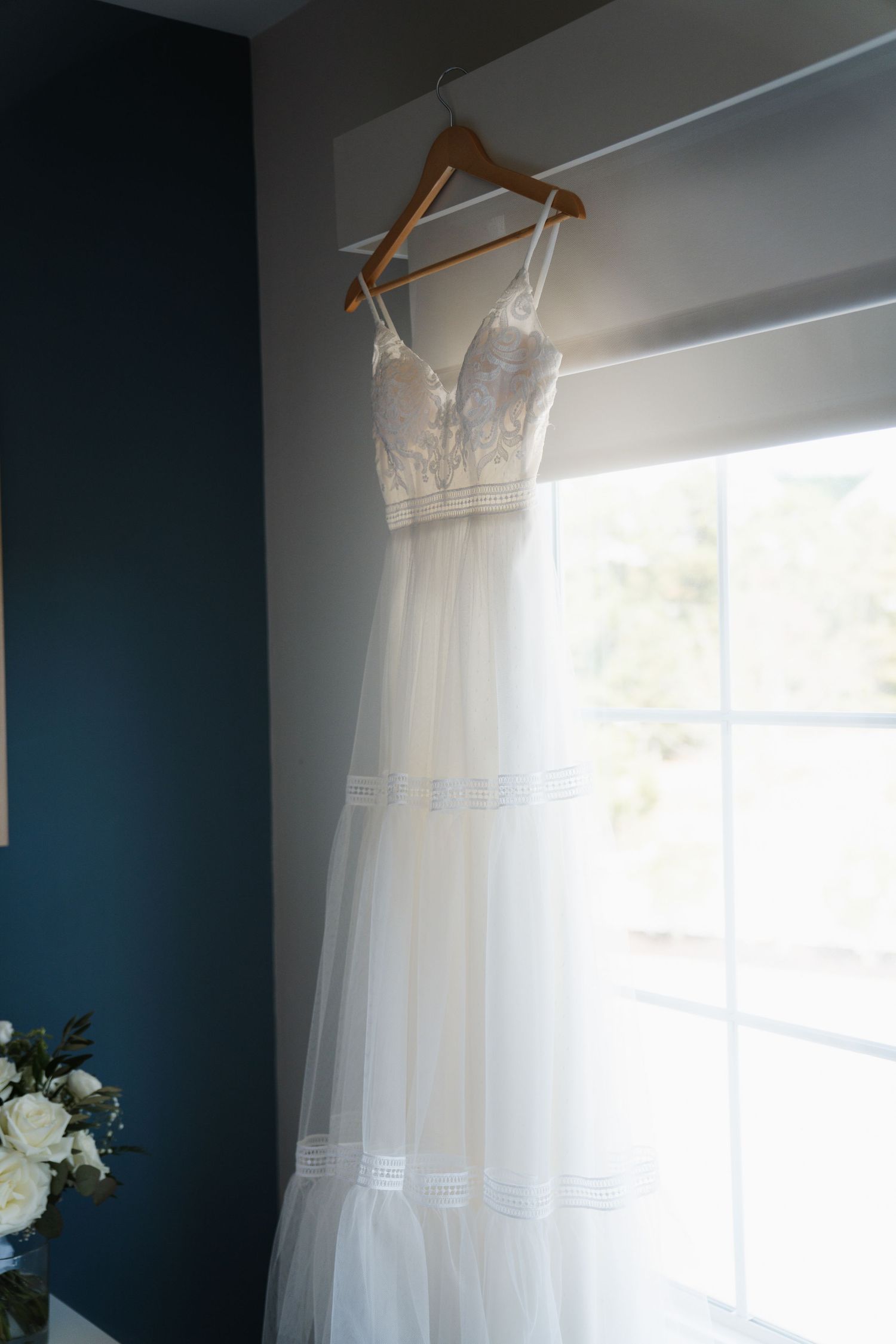 A white wedding dress hangs by a window with natural light streaming in against a dark blue wall.