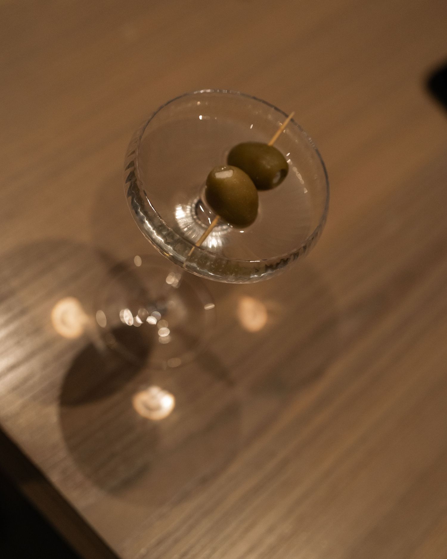 A martini glass with two olives photographed from above on a wooden surface.