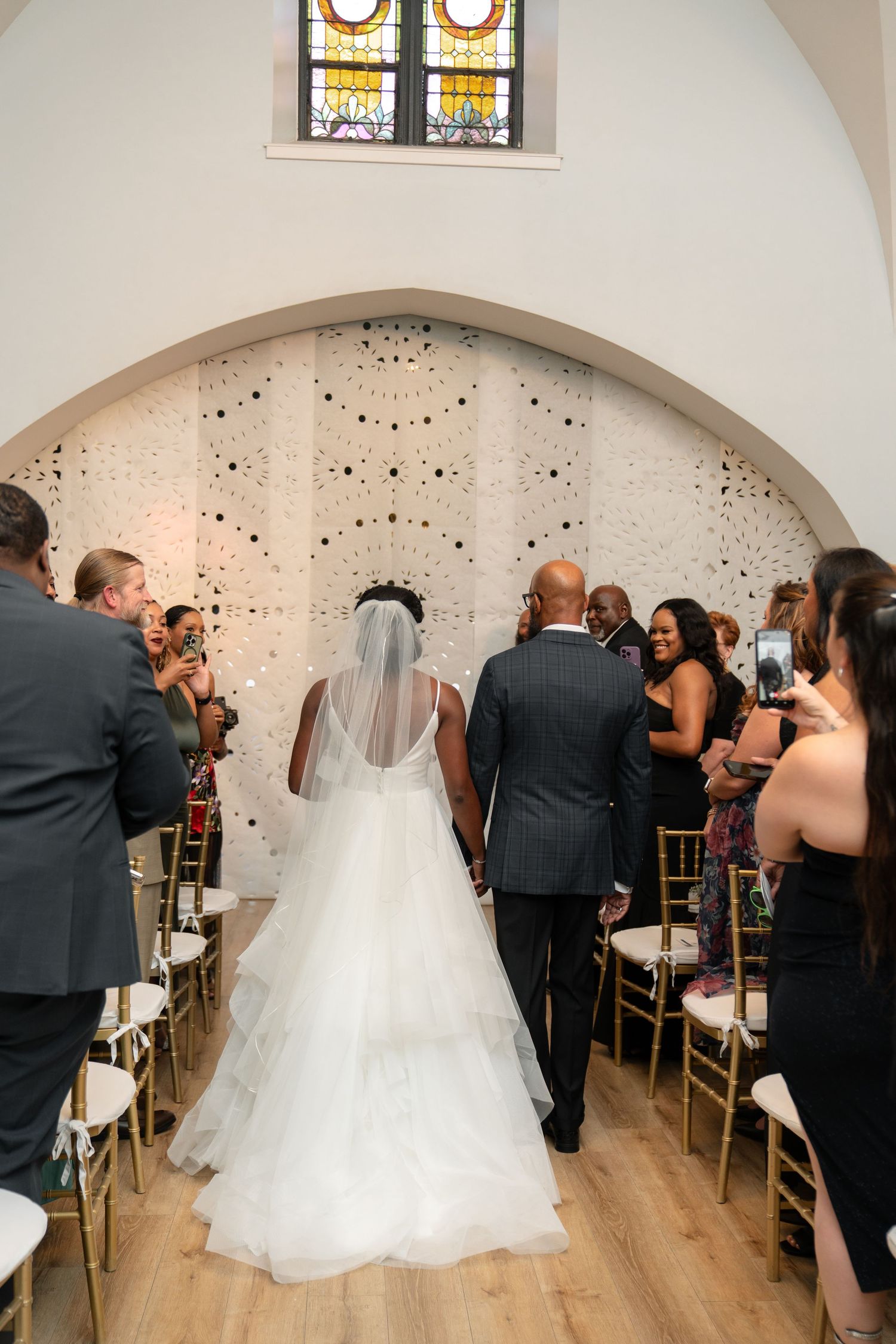 Wedding ceremony in progress inside a bright venue with stained glass windows and wooden floors.