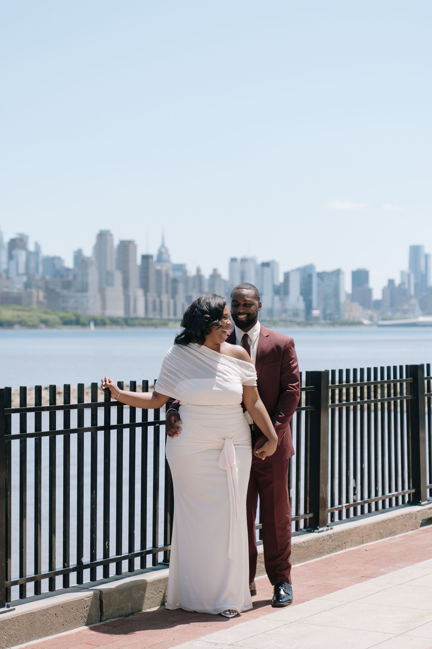 Couple in wedding attire posing by waterfront railing with New York City skyline in background on sunny day.