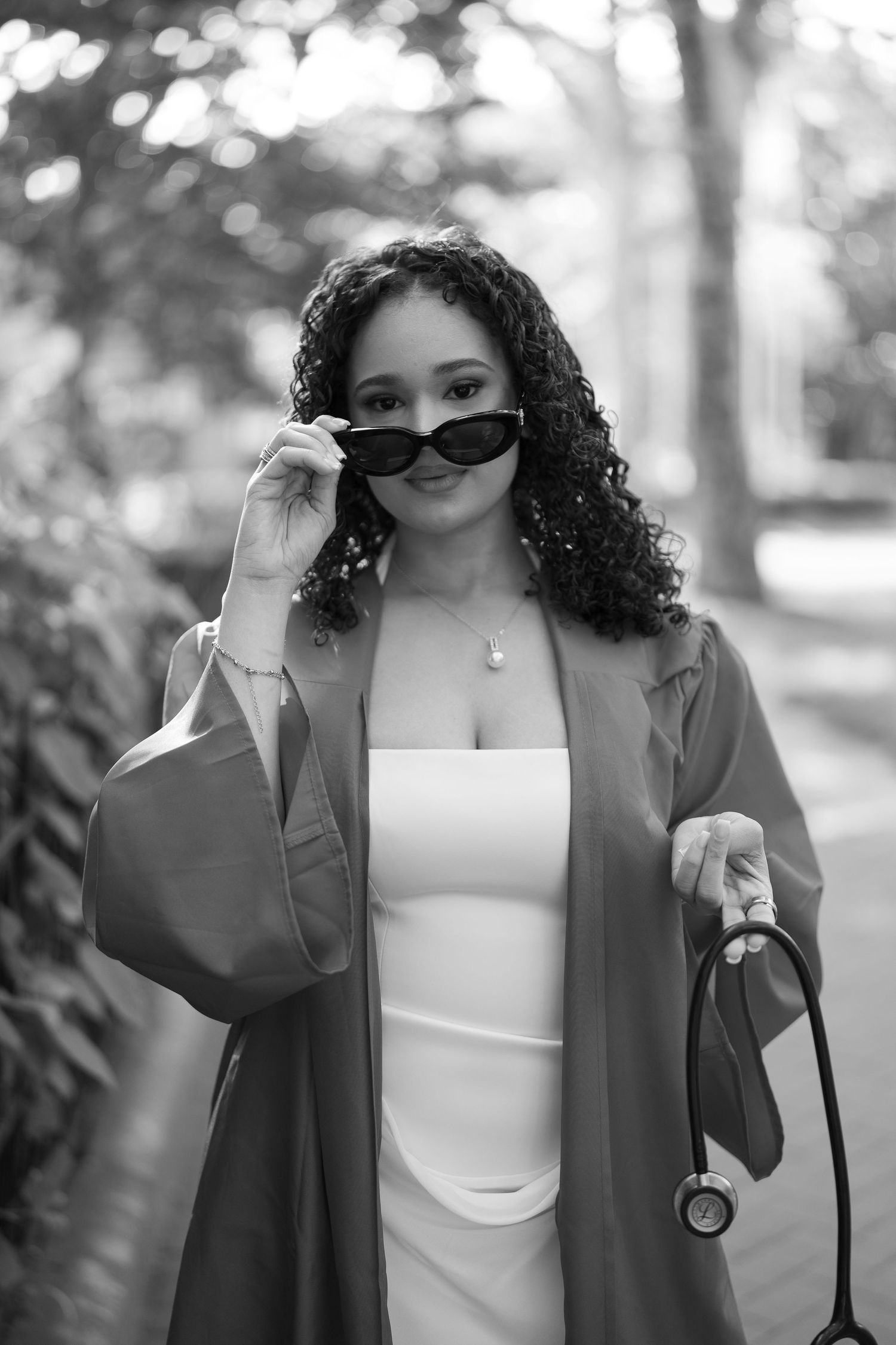 Curly-haired woman in graduation gown peers over sunglasses in outdoor black and white photo.