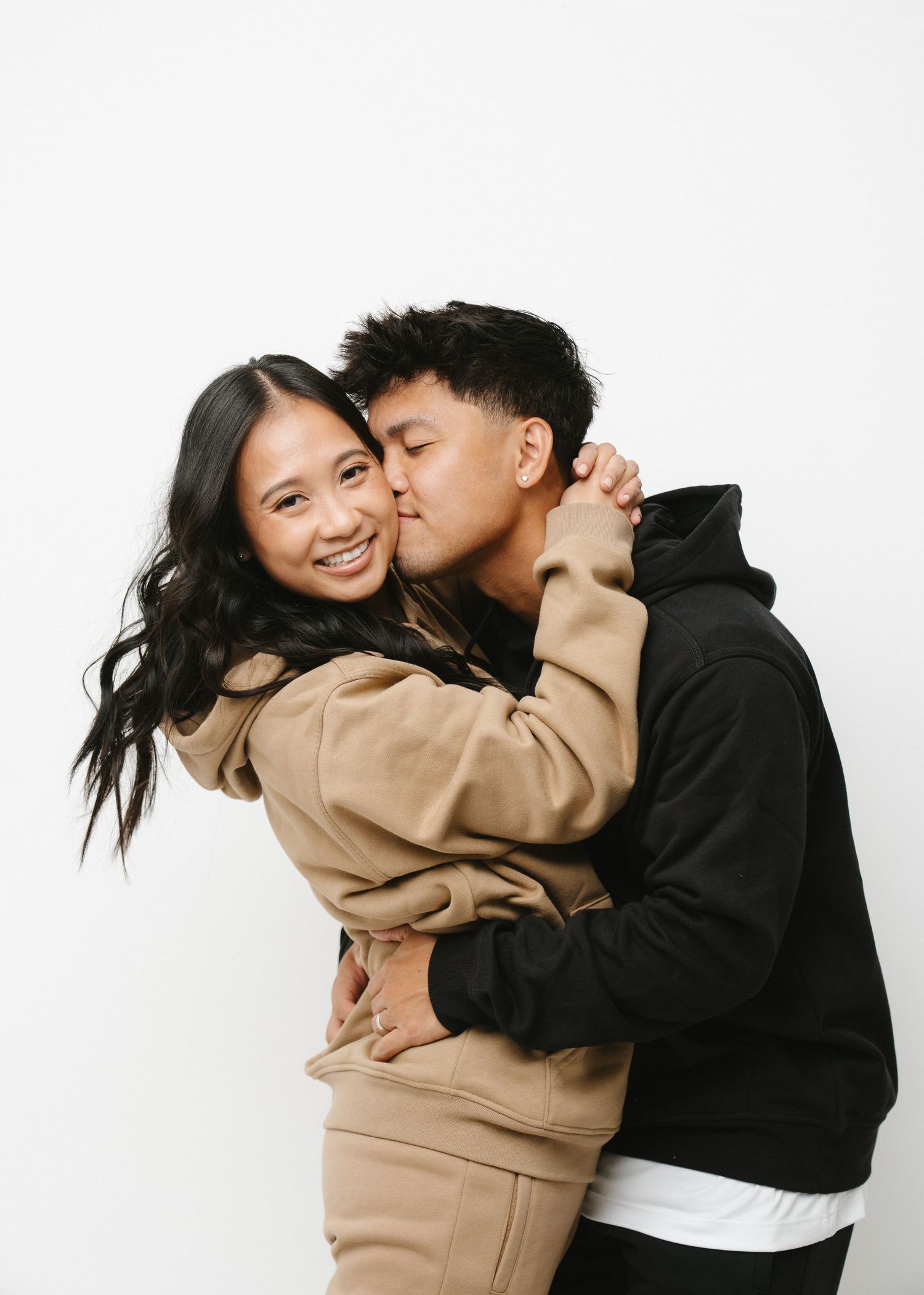 A couple shares playful moments wearing black and beige hoodies against a white studio background.