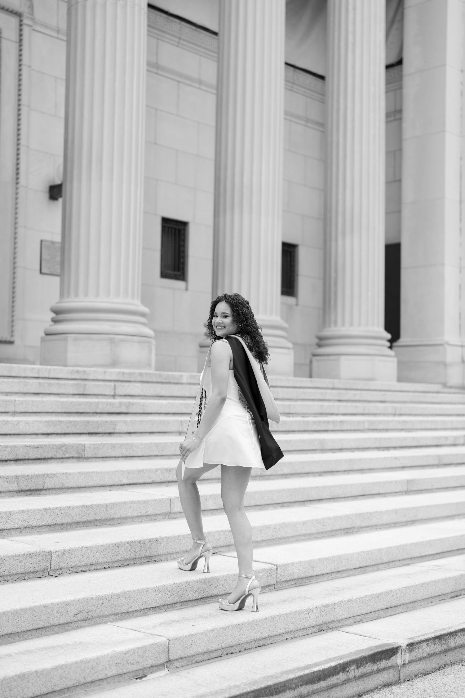 Black and white photo of smiling graduate in white dress on grand building steps.