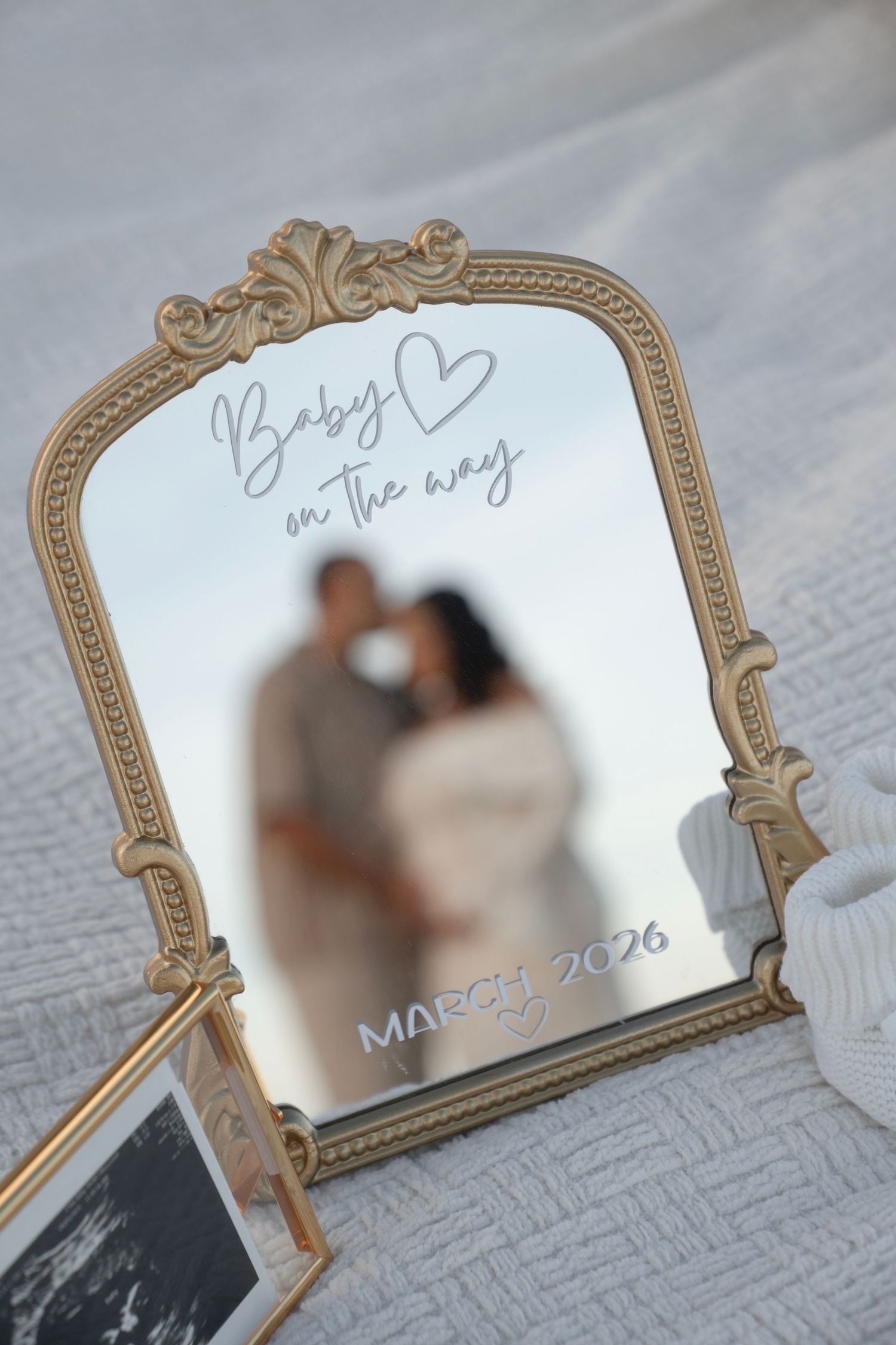 Gold ornate mirror with Baby on the Way March 2026 announcement, blurred couple reflection, ultrasound photo, pregnancy reveal.