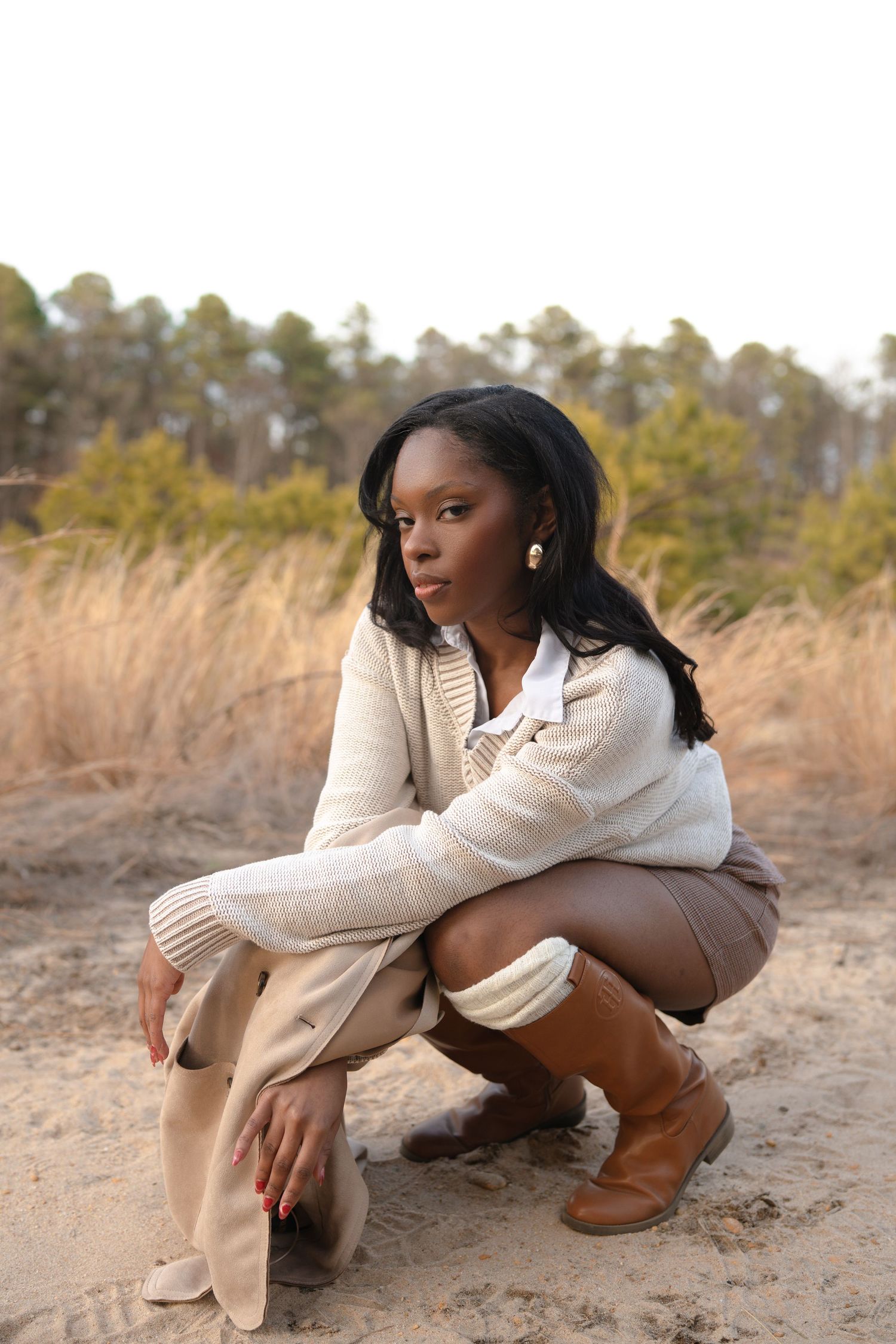 Model in white knit sweater crouches gracefully in outdoor nature setting.