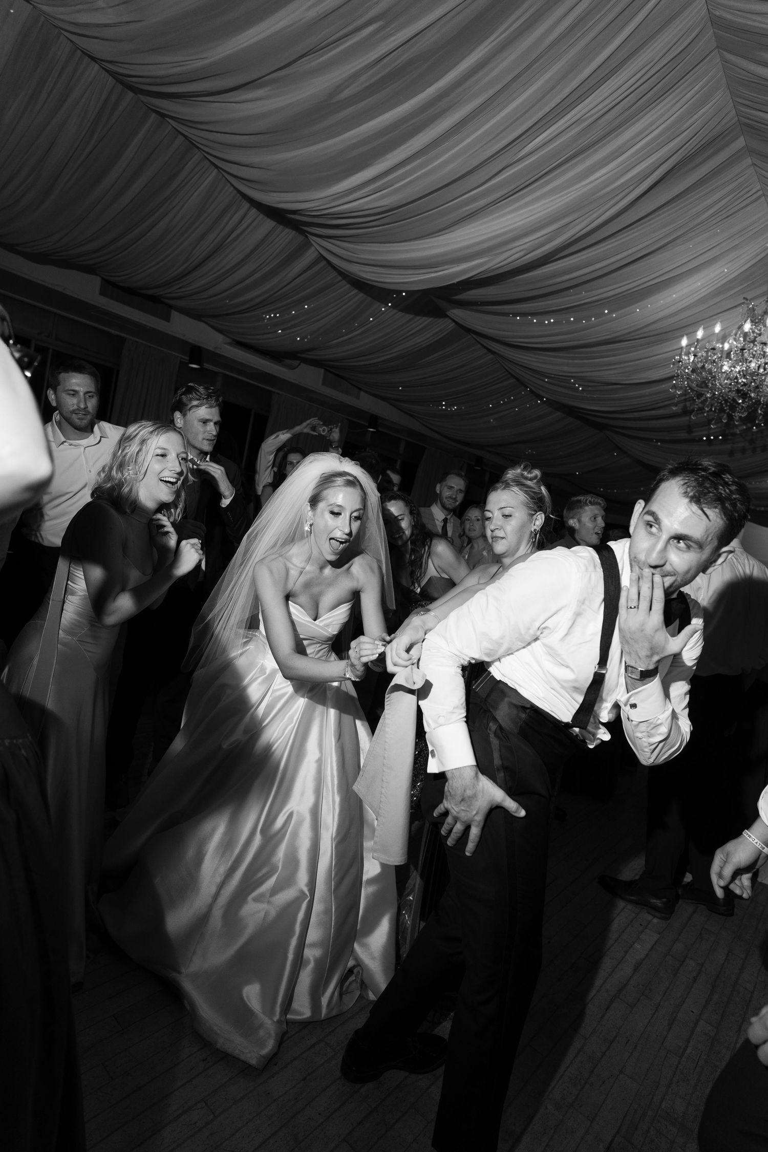 Bride and groom dancing joyfully with guests on the wedding reception dance floor in black and white.