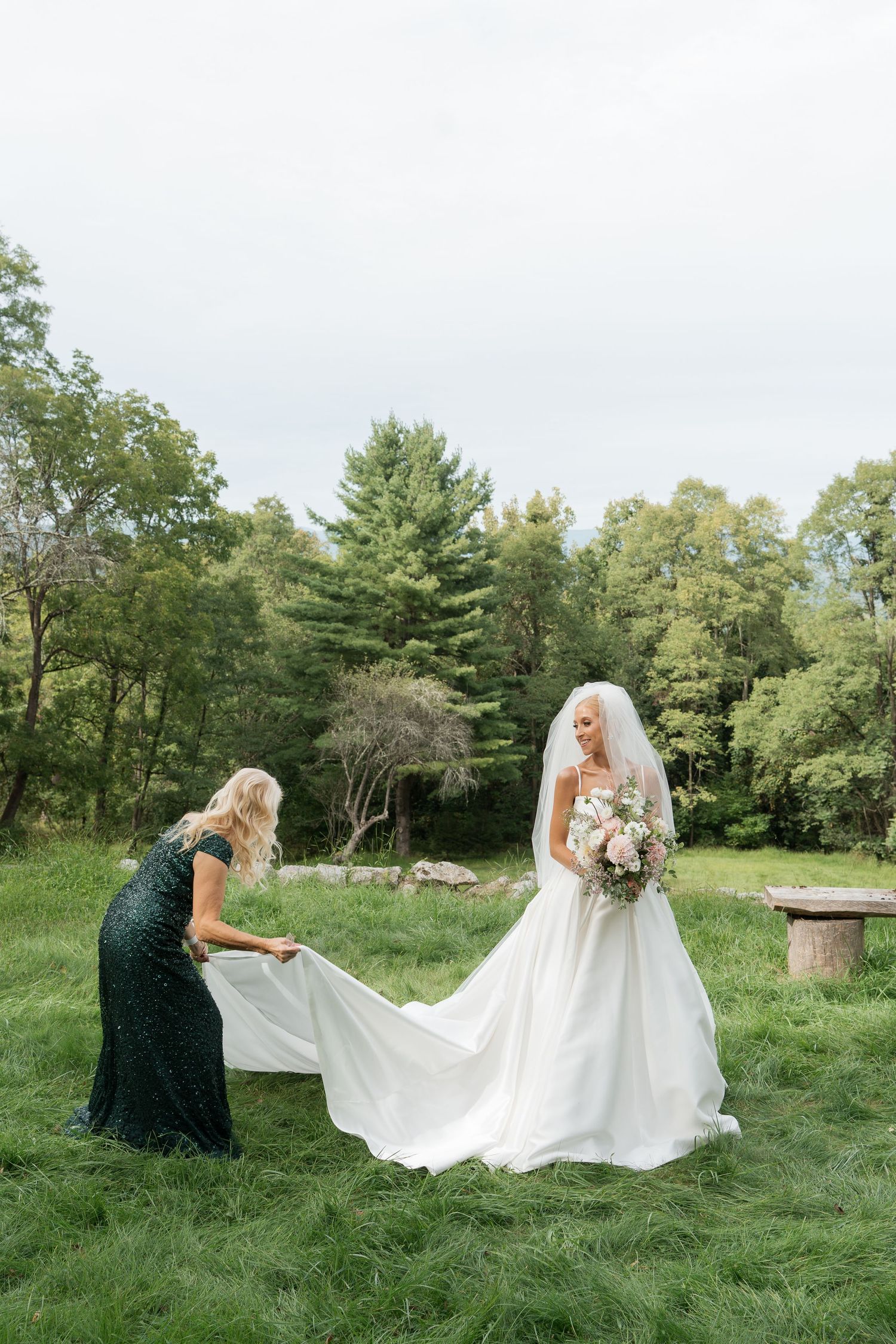 Bridesmaid in green sequin dress adjusting the train of a bride holding a bouquet on a lush green lawn.