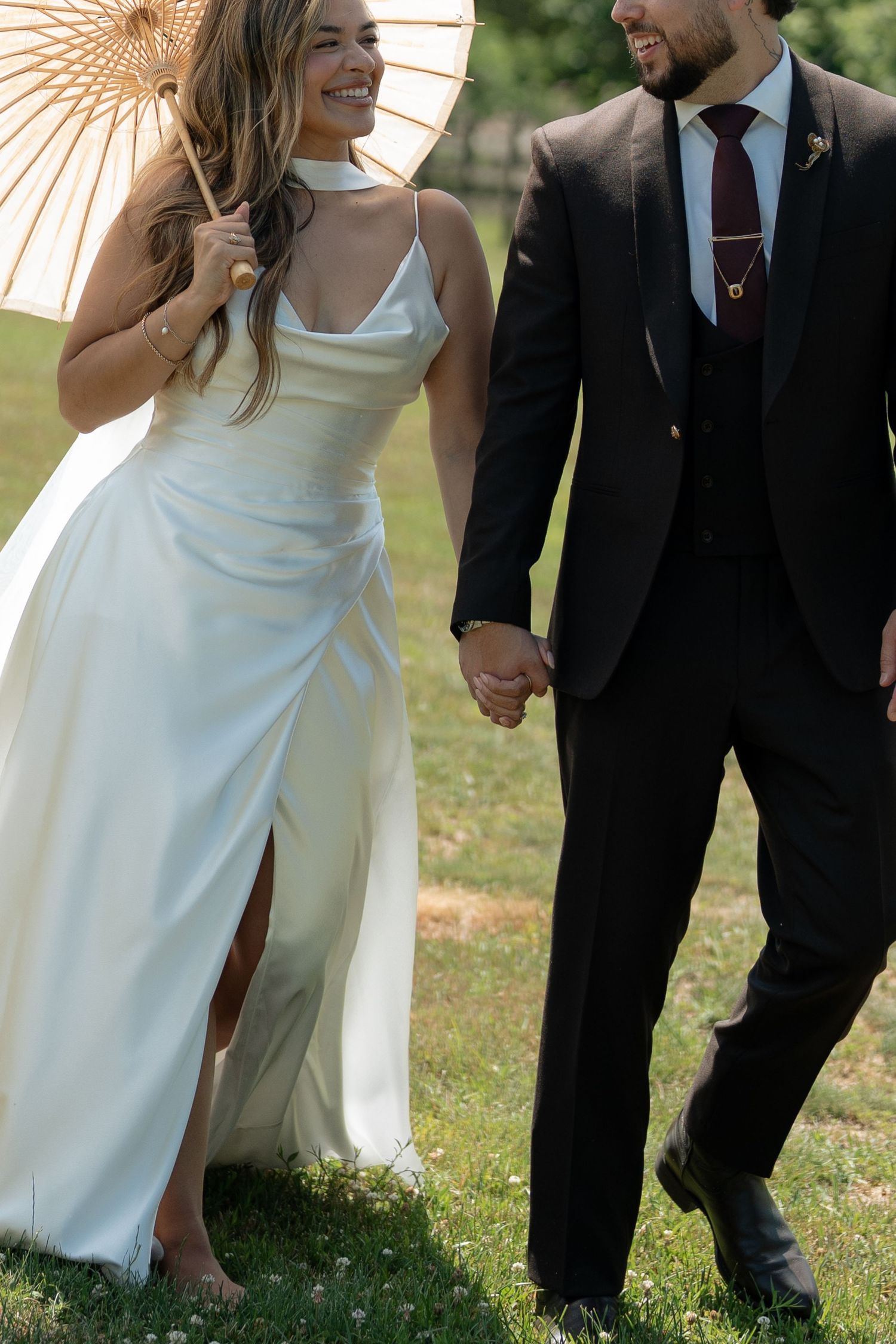 A smiling bride in a white satin gown holds a parasol while walking hand in hand with her groom outdoors.