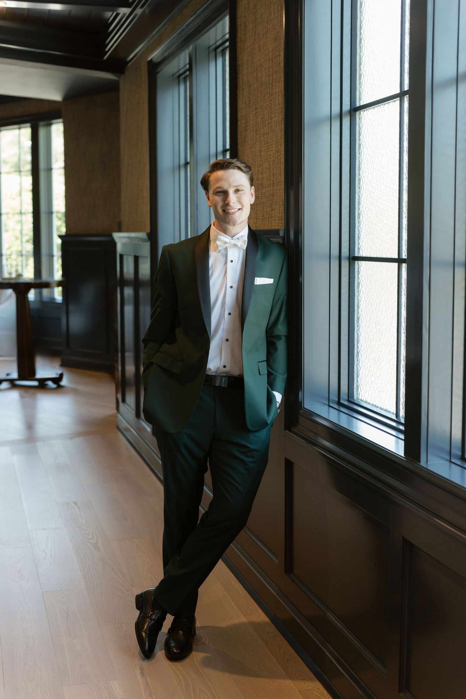 Smiling man in green suit leaning against wall by windows in elegant interior.