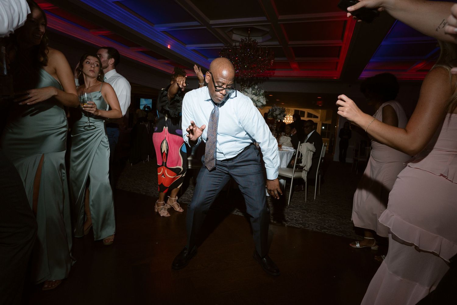 Man in suit dancing energetically at a wedding reception party with guests in formal attire in background.