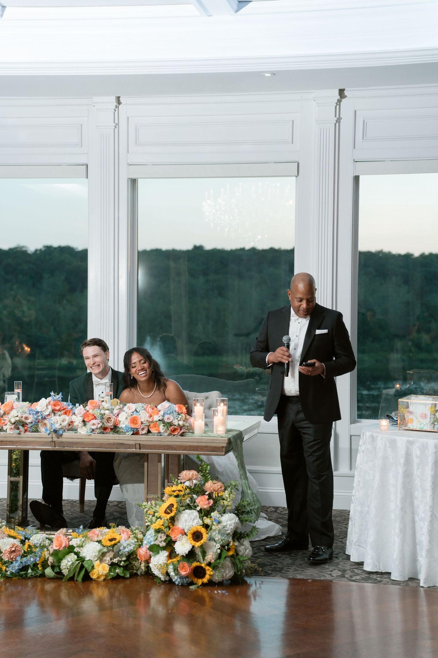 Father delivers wedding toast as bride and groom laugh at sweetheart table adorned with sunflower arrangements.