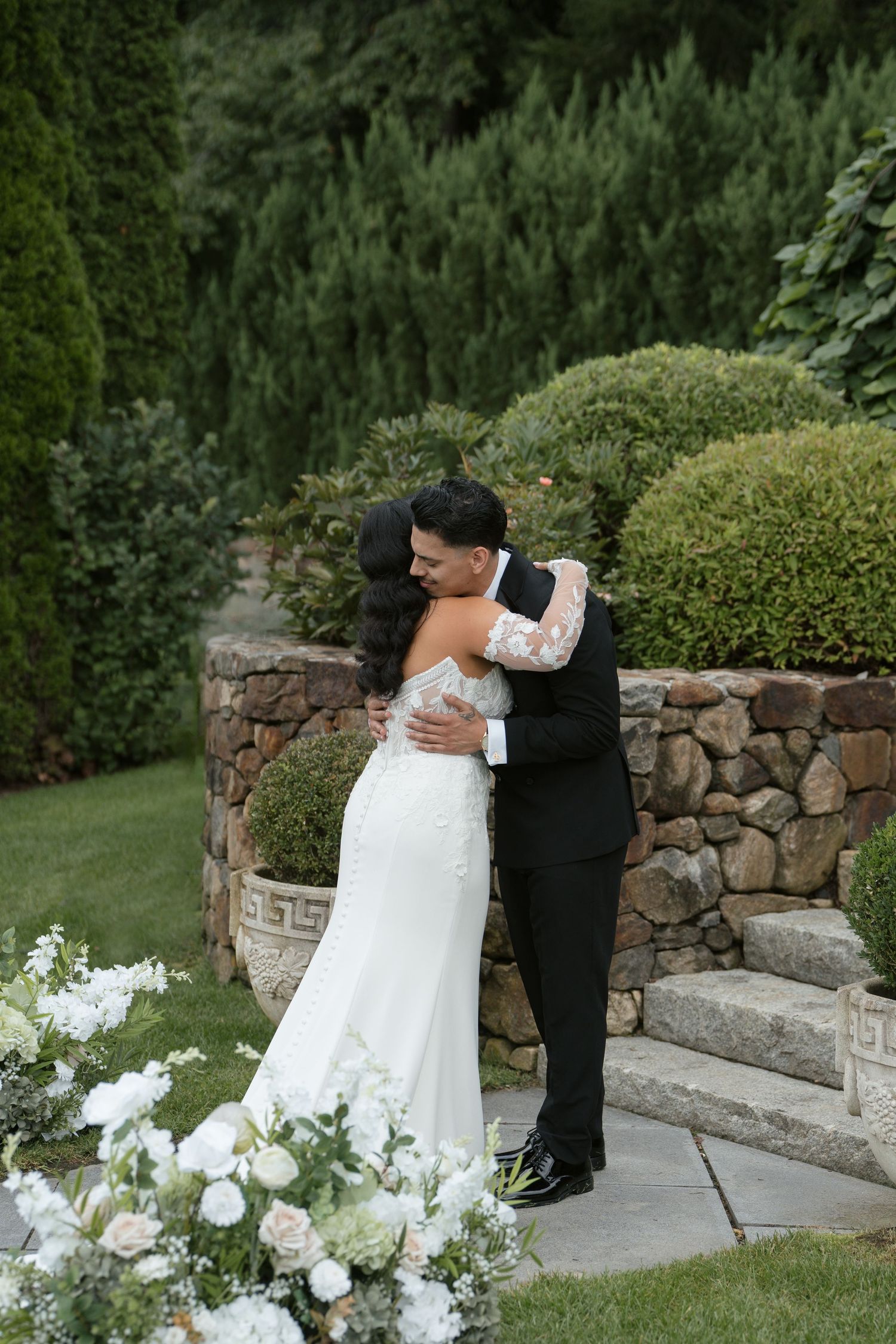 Bride and groom share a heartfelt embrace in a lush garden ceremony, surrounded by elegant white floral arrangements.