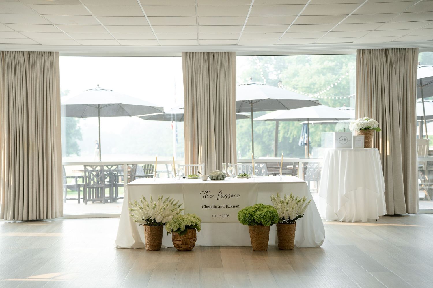 Elegant wedding reception head table with white linen, floral arrangements, and large windows overlooking outdoor patio.