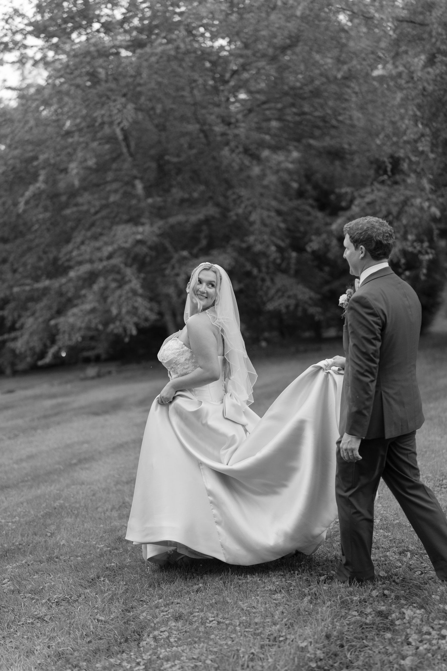 Black and white photo of smiling bride in veil as groom carries her satin train outdoors.