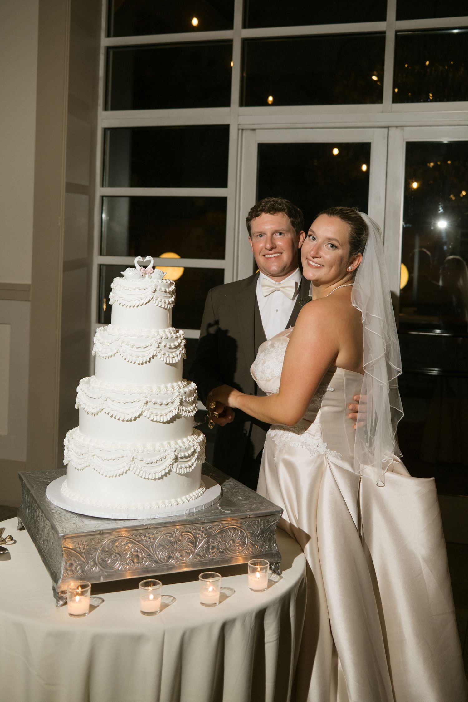 Bride and groom smiling while cutting their elegant four-tier white wedding cake adorned with pearl details and candles.