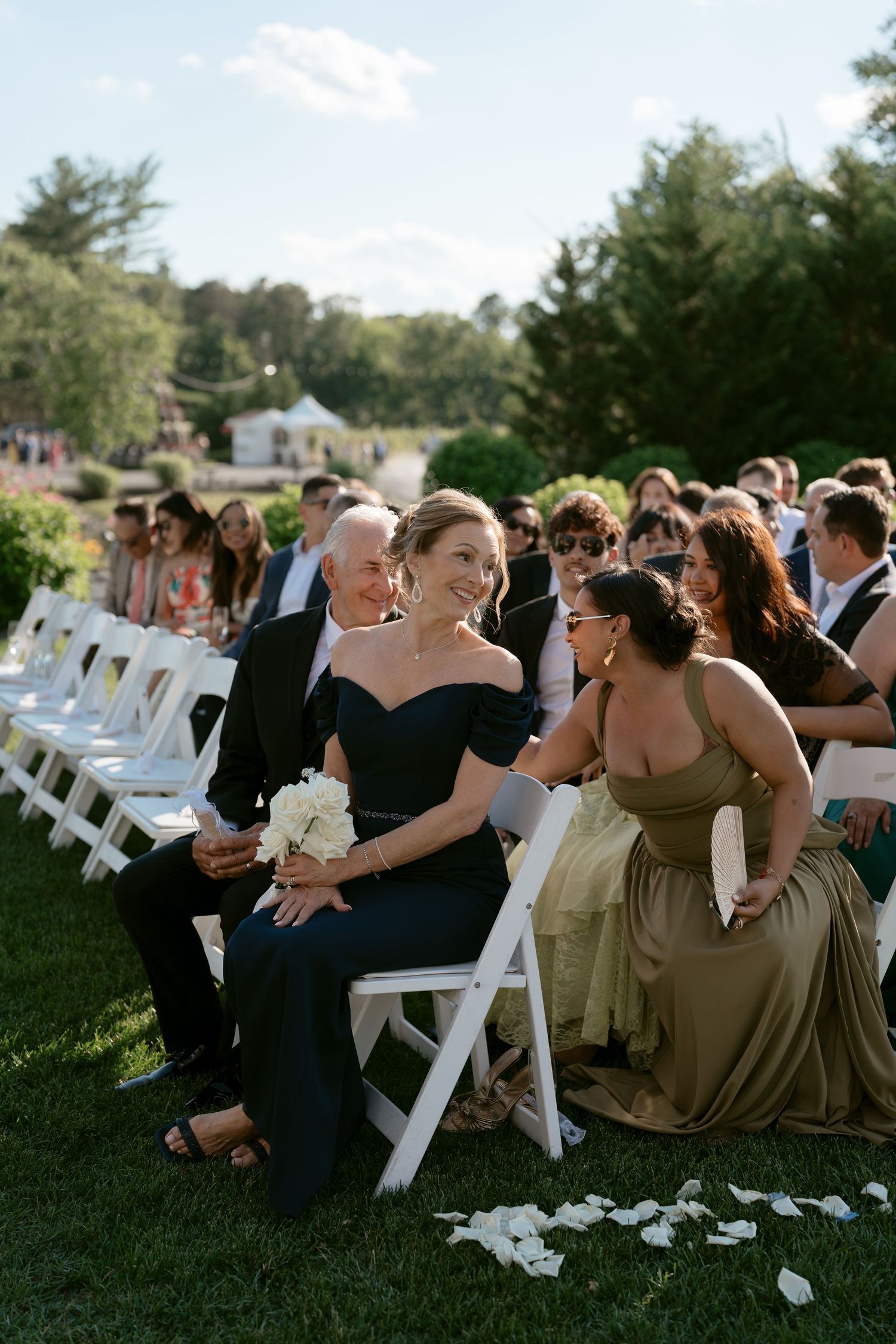 Guests smile and laugh during an outdoor wedding ceremony, seated on white chairs on a sunny green lawn.