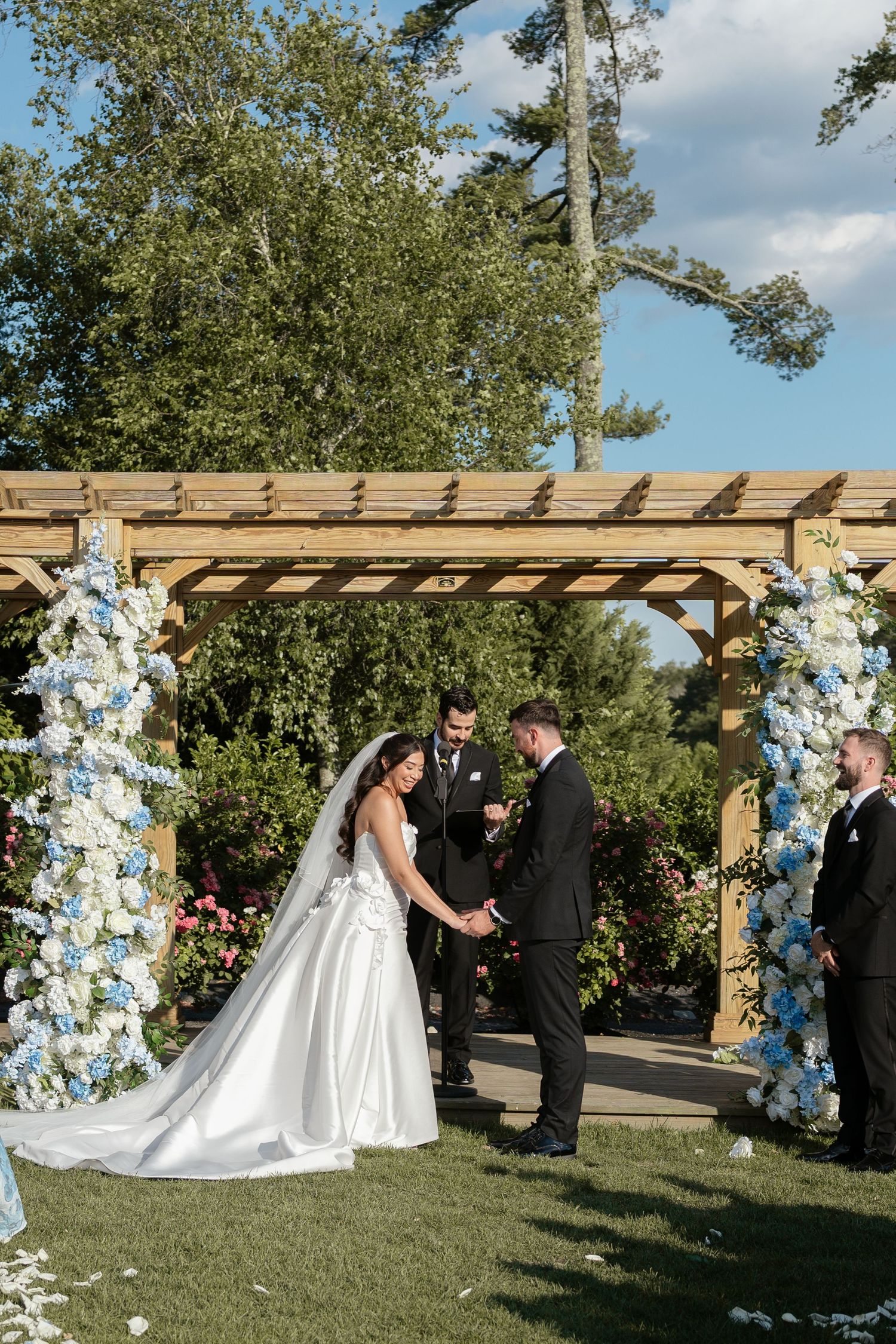 Bride and groom exchanging vows under wooden pergola with blue and white floral arch.