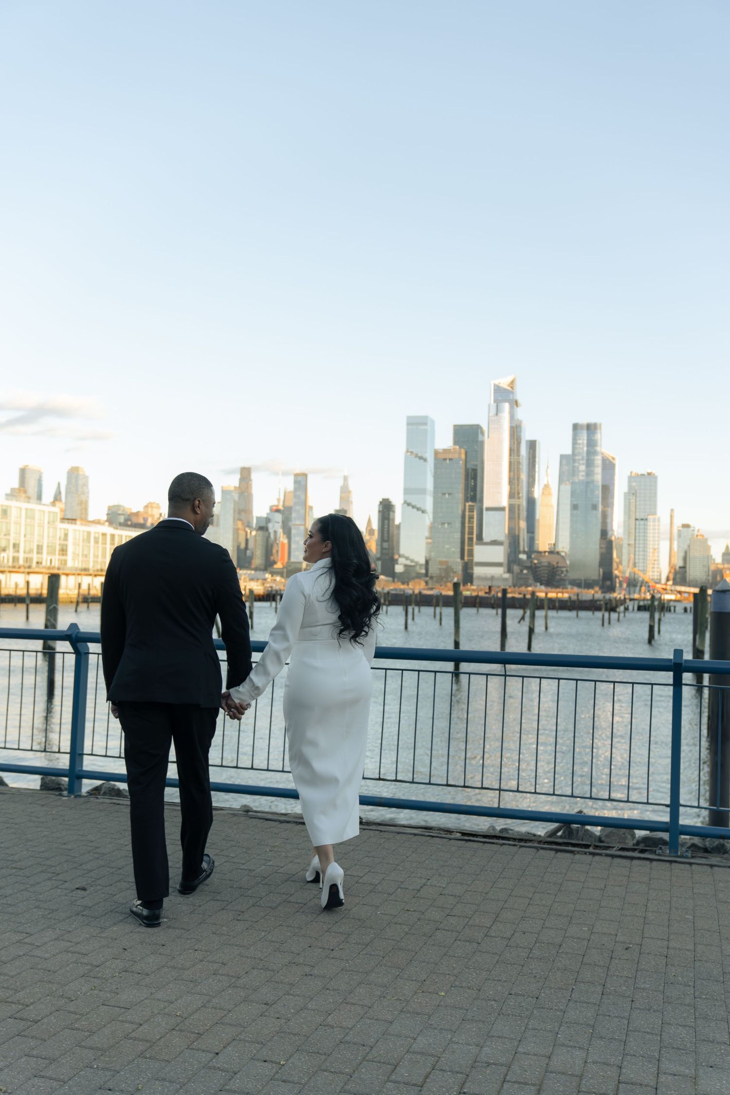 A couple walks along a waterfront with the New York City skyline in the background.