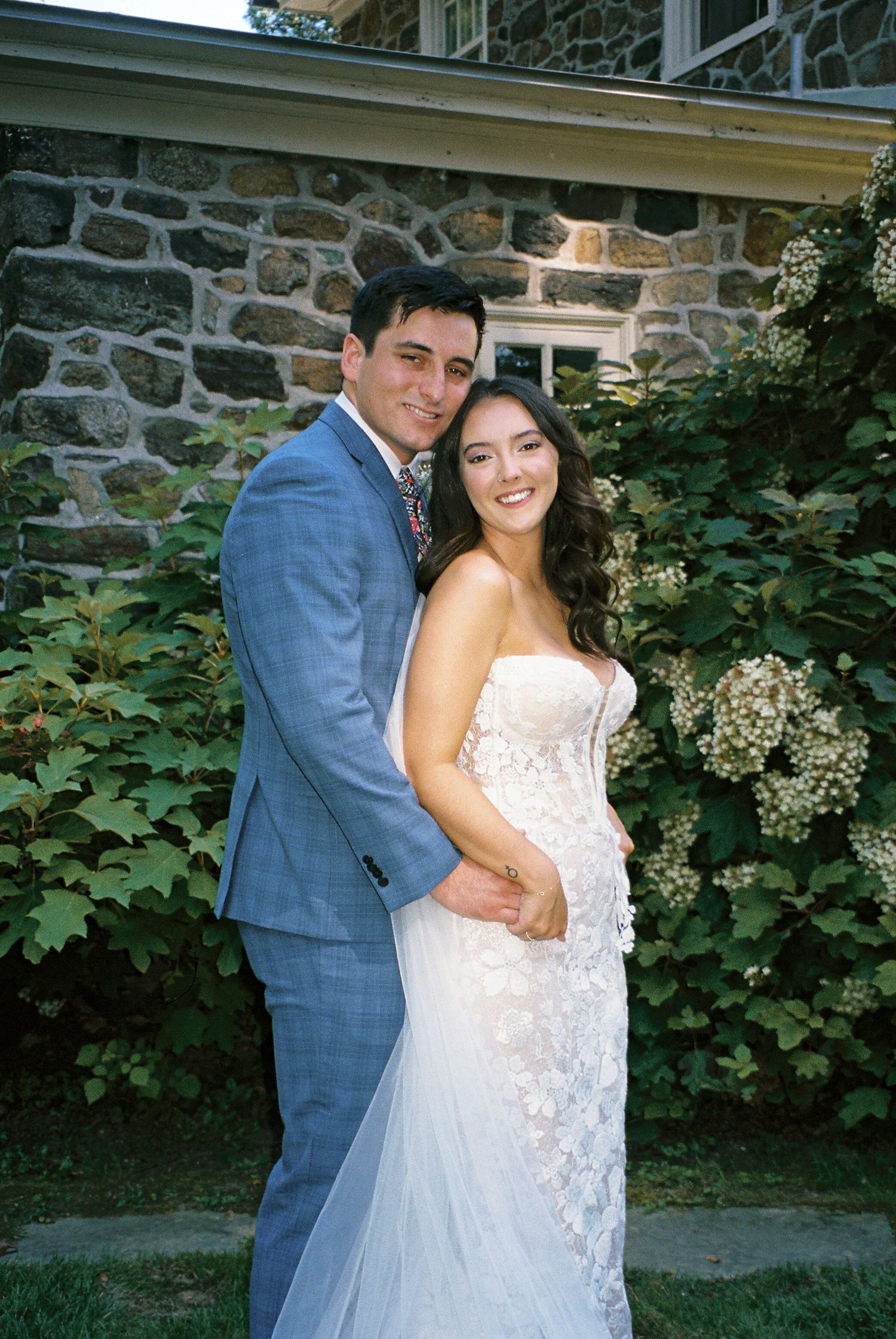 A romantic wedding photoshoot featuring a couple embracing against a stone wall backdrop with greenery.
