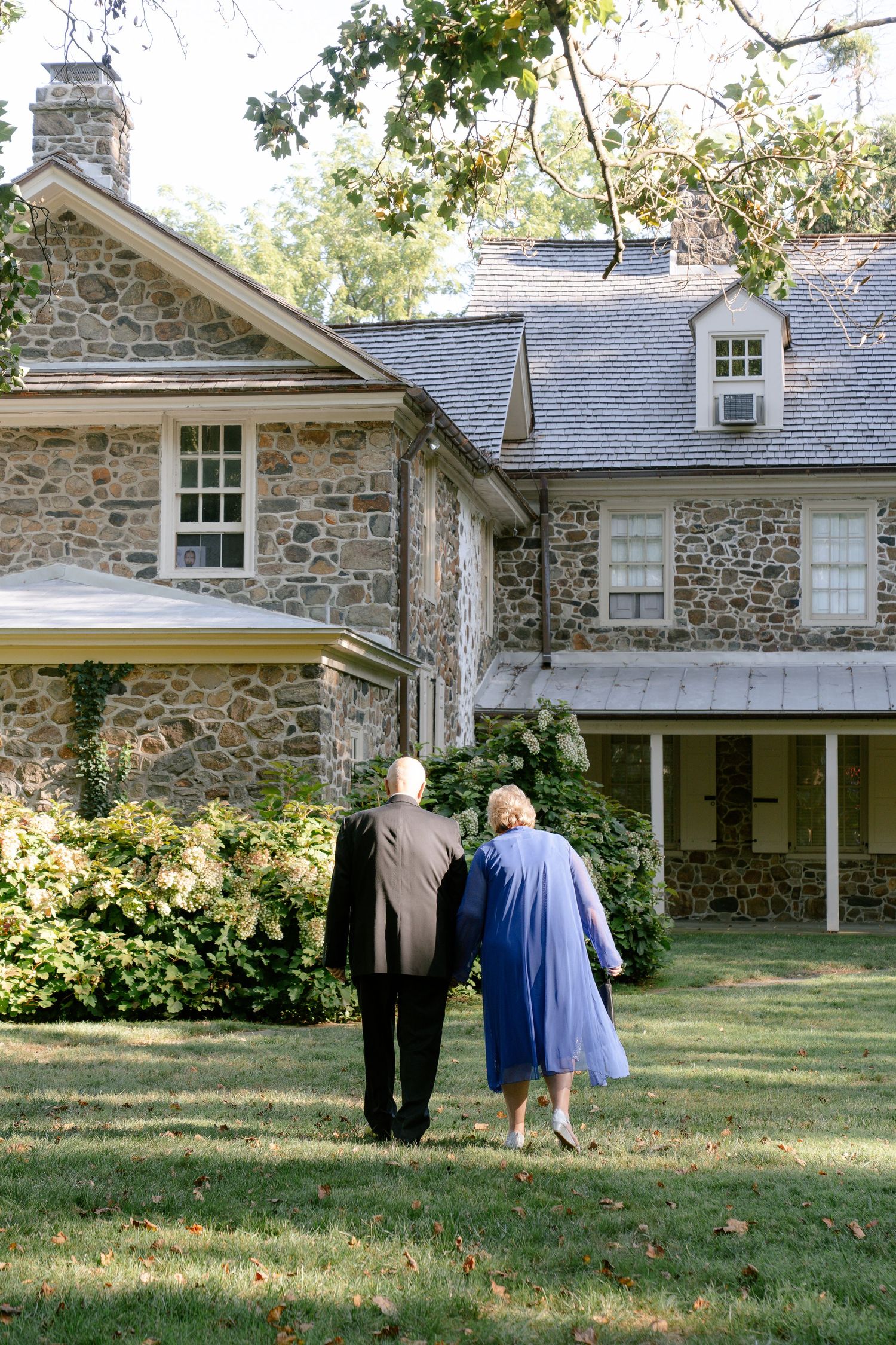 Historic stone house with white trim windows and wraparound porch in a garden setting.