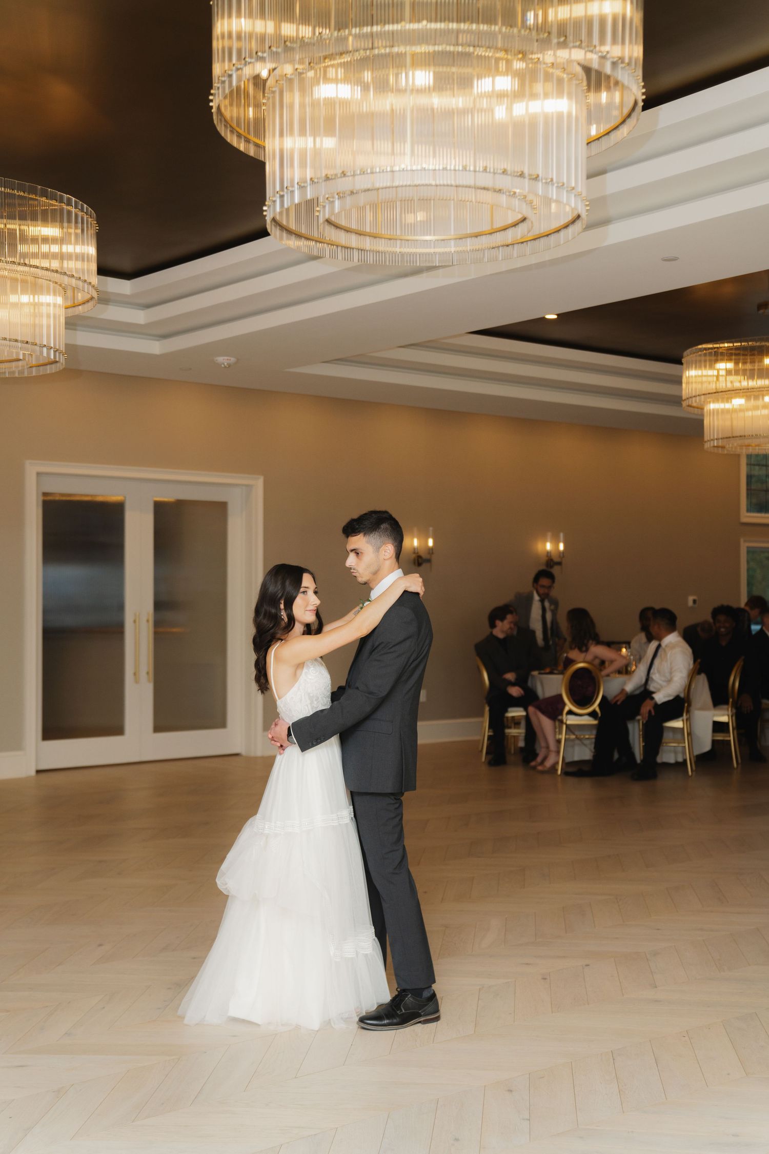 A couple shares their first dance in an elegant ballroom with crystal chandeliers and white decor.