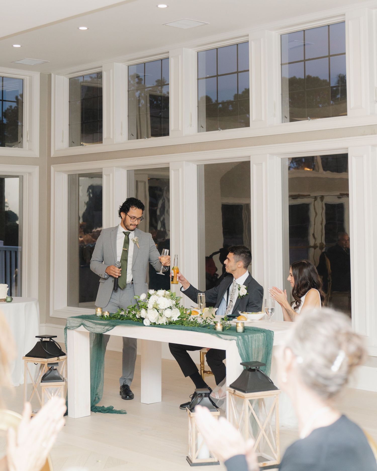A wedding reception scene with people giving toasts at a white-walled venue with large windows and elegant floral decor.