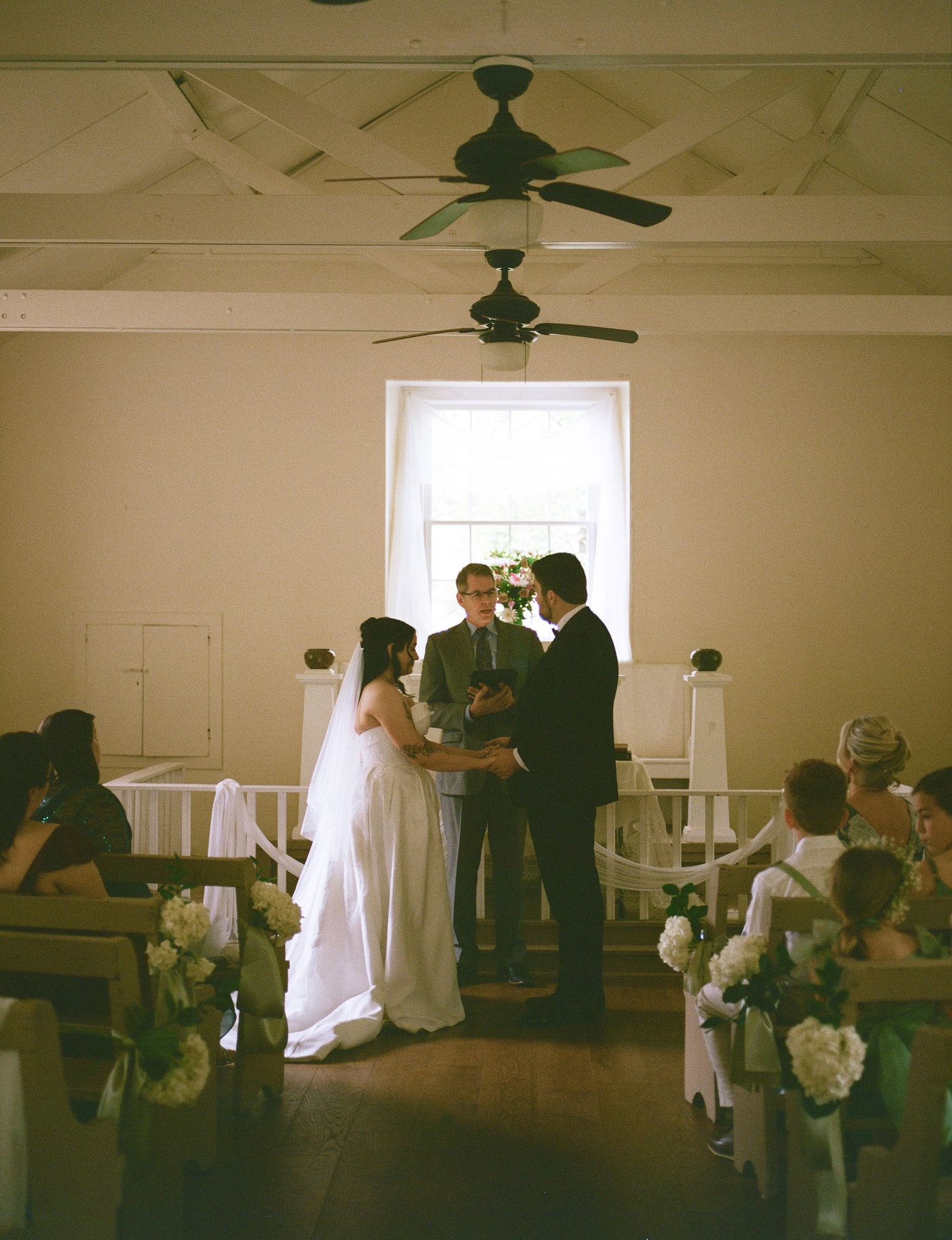 Bride and groom exchange vows at rustic chapel altar with officiant, white pews, and ceiling fan overhead.