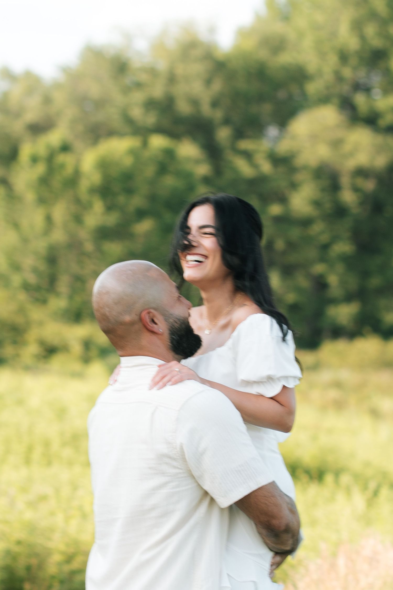 A couple in white outfits share a joyful moment together in a sunlit meadow with green trees in the background.