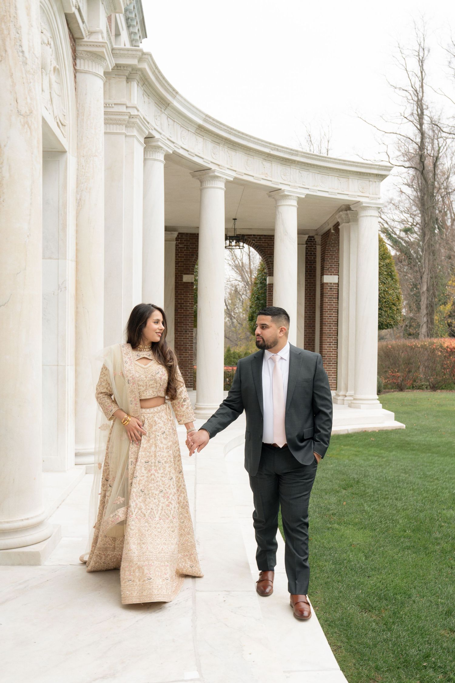 A couple shares romantic moments along a white columned colonnade walkway with green lawn in the background.