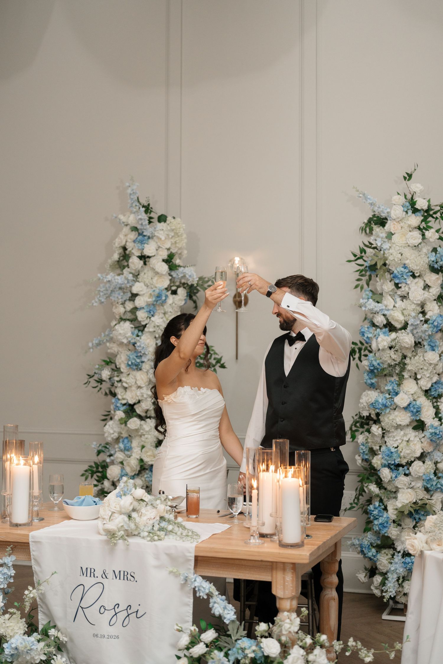 Mr. and Mrs. Rossi toast with champagne at sweetheart table surrounded by blue and white florals.