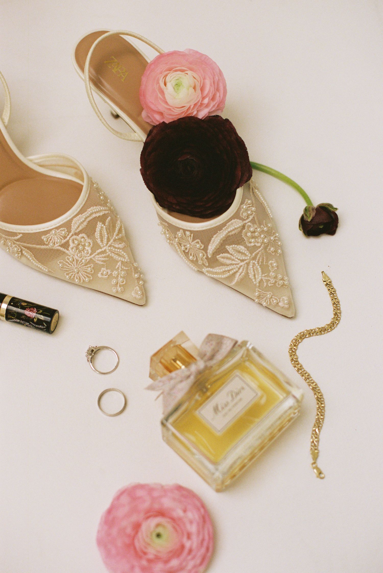Elegant bridal flat shoes with floral embroidery, Miss Dior perfume, gold bracelet, rings, and pink ranunculus flowers.