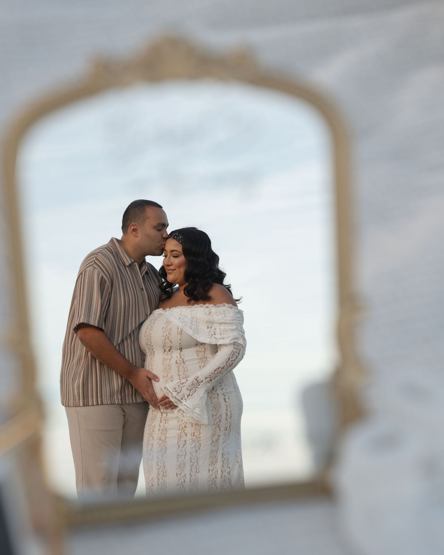 Couple sharing a tender moment reflected in an ornate gold mirror, woman in white lace maternity gown during outdoor photoshoot.