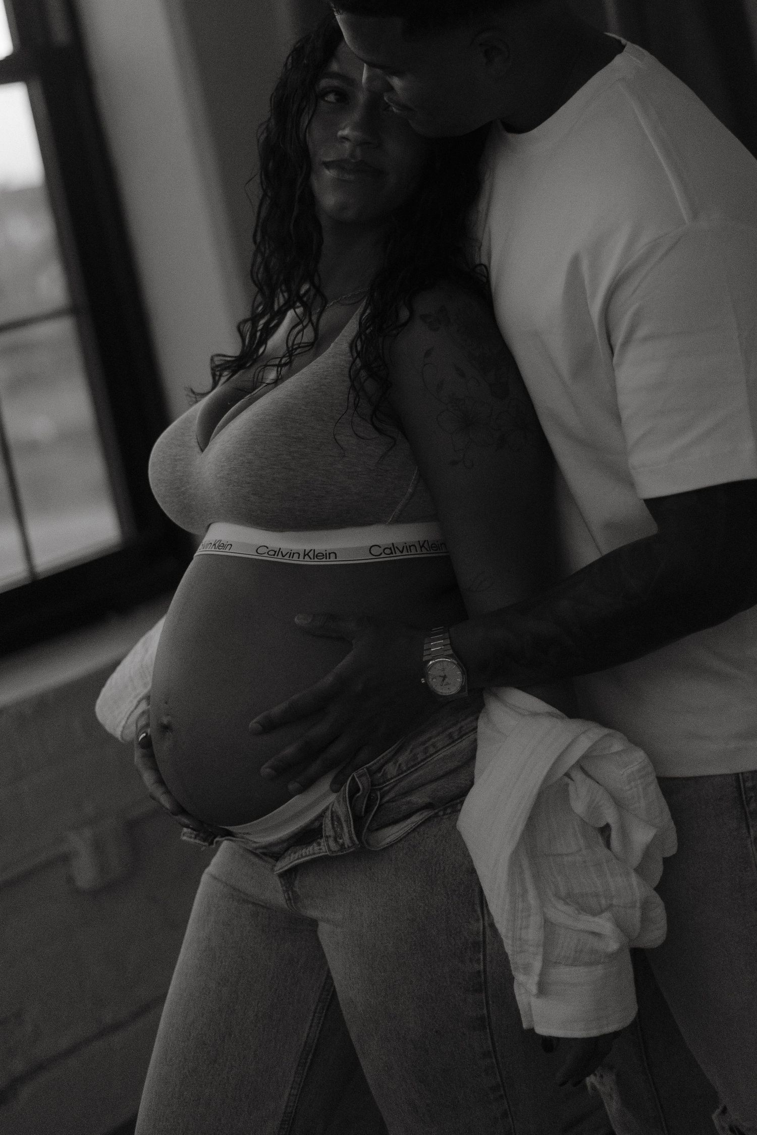 Black and white maternity photo of a pregnant woman in Calvin Klein bra and jeans, embraced by her partner.