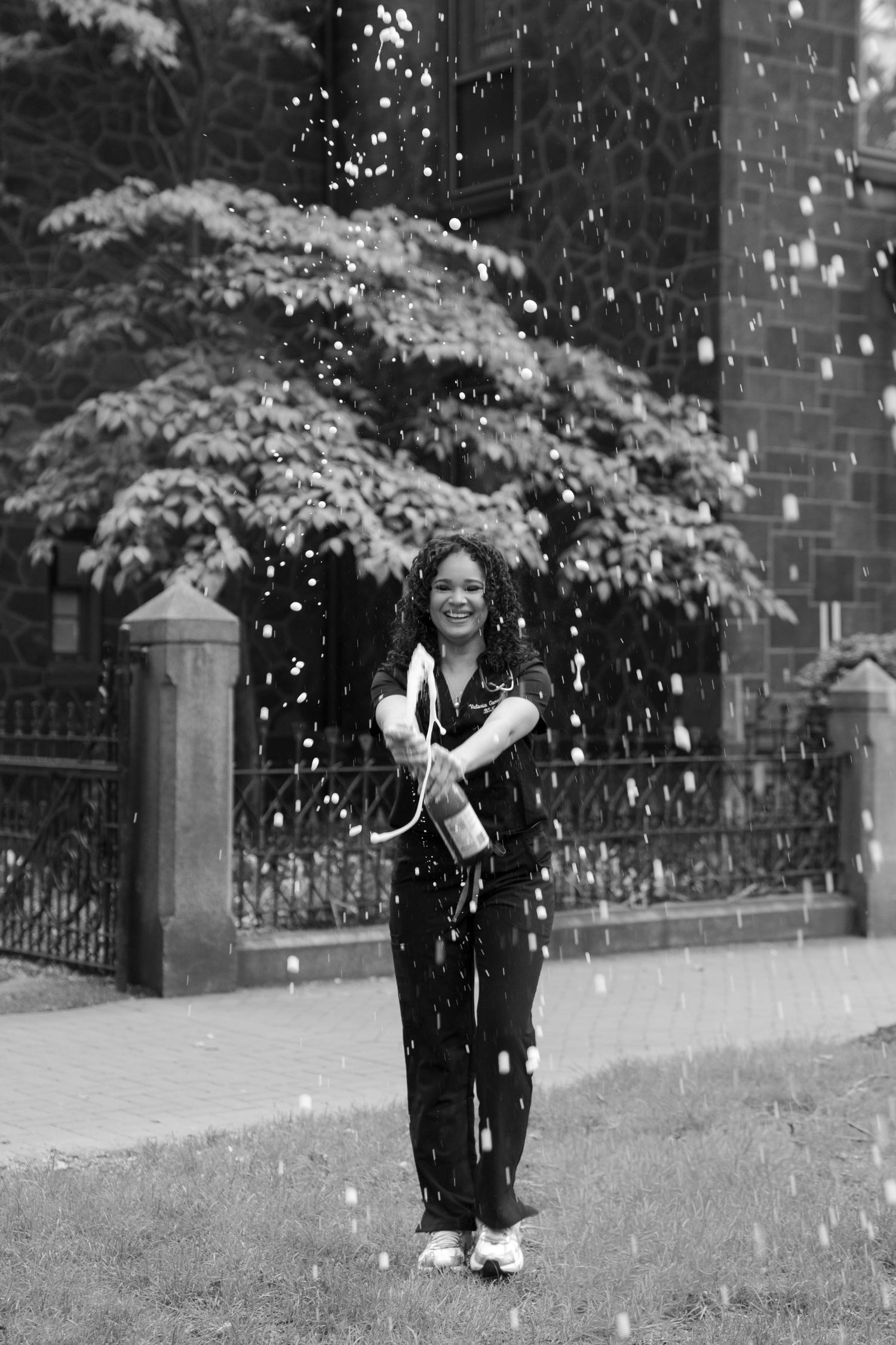 Black and white photo of graduate celebrating with confetti outdoors in scrubs.