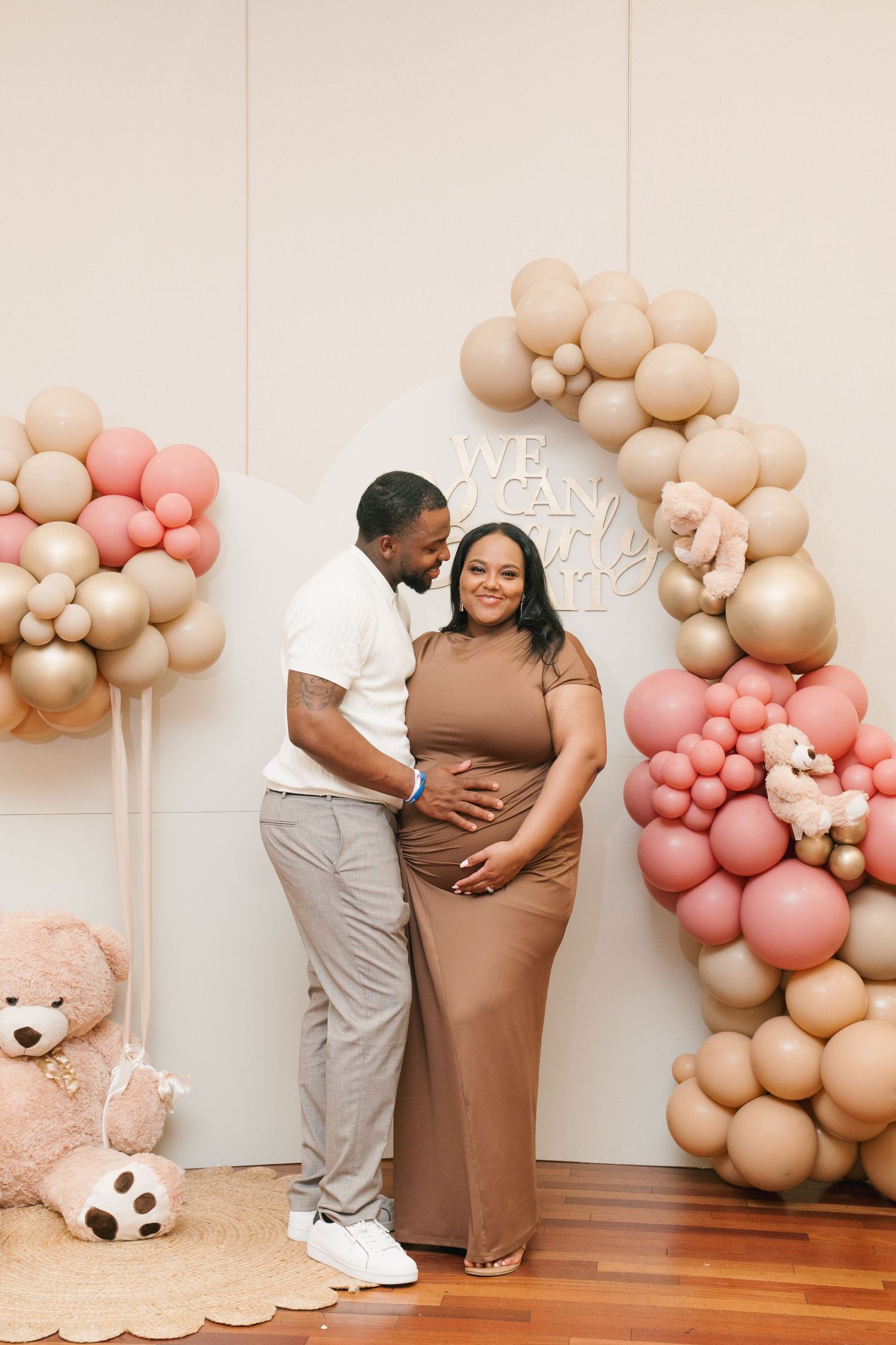 A pregnancy announcement photoshoot featuring balloon decorations in soft pink and beige tones against a white wall.