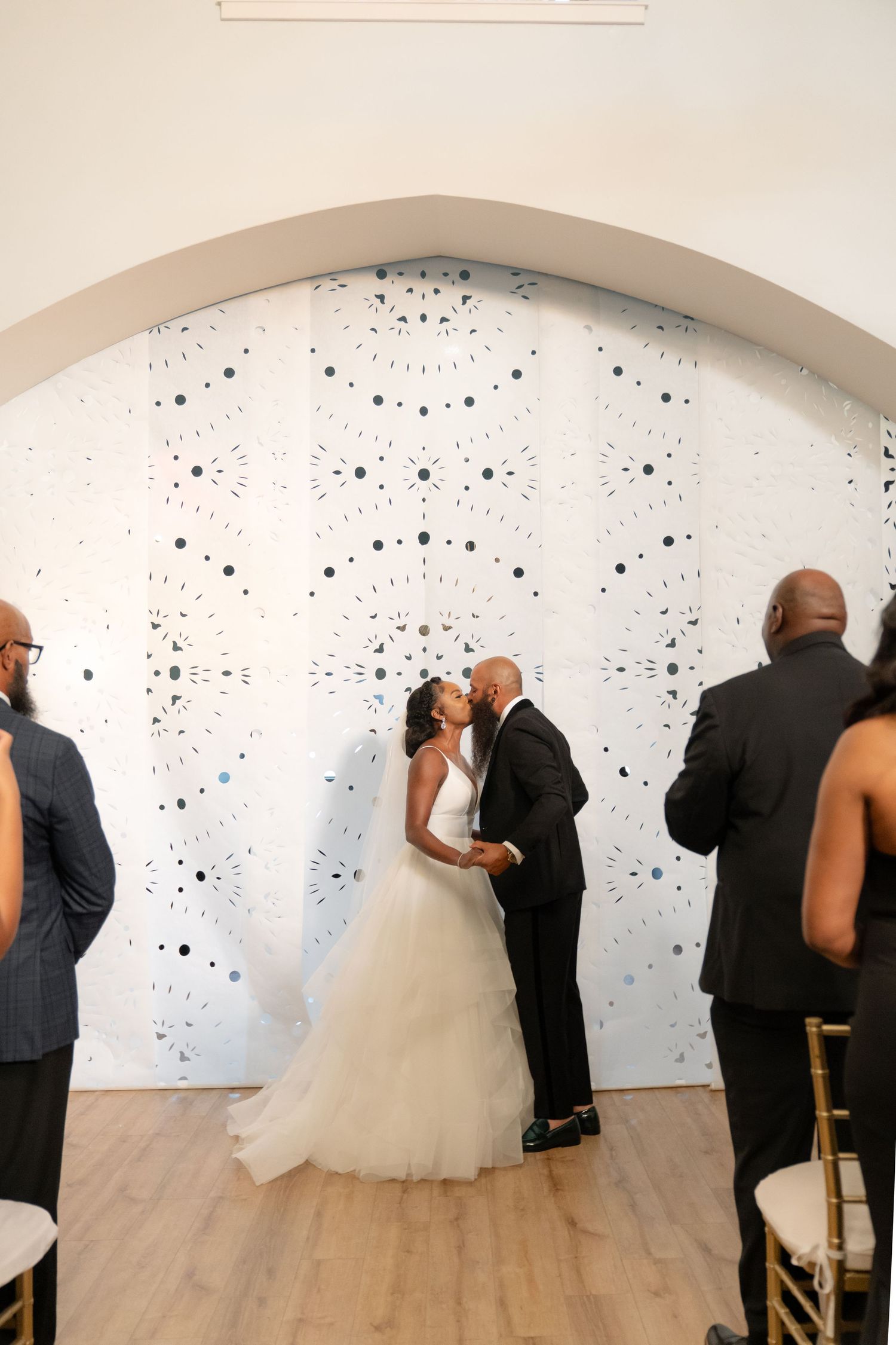 Wedding couple poses for photos against a white wall with artistic black dot pattern.