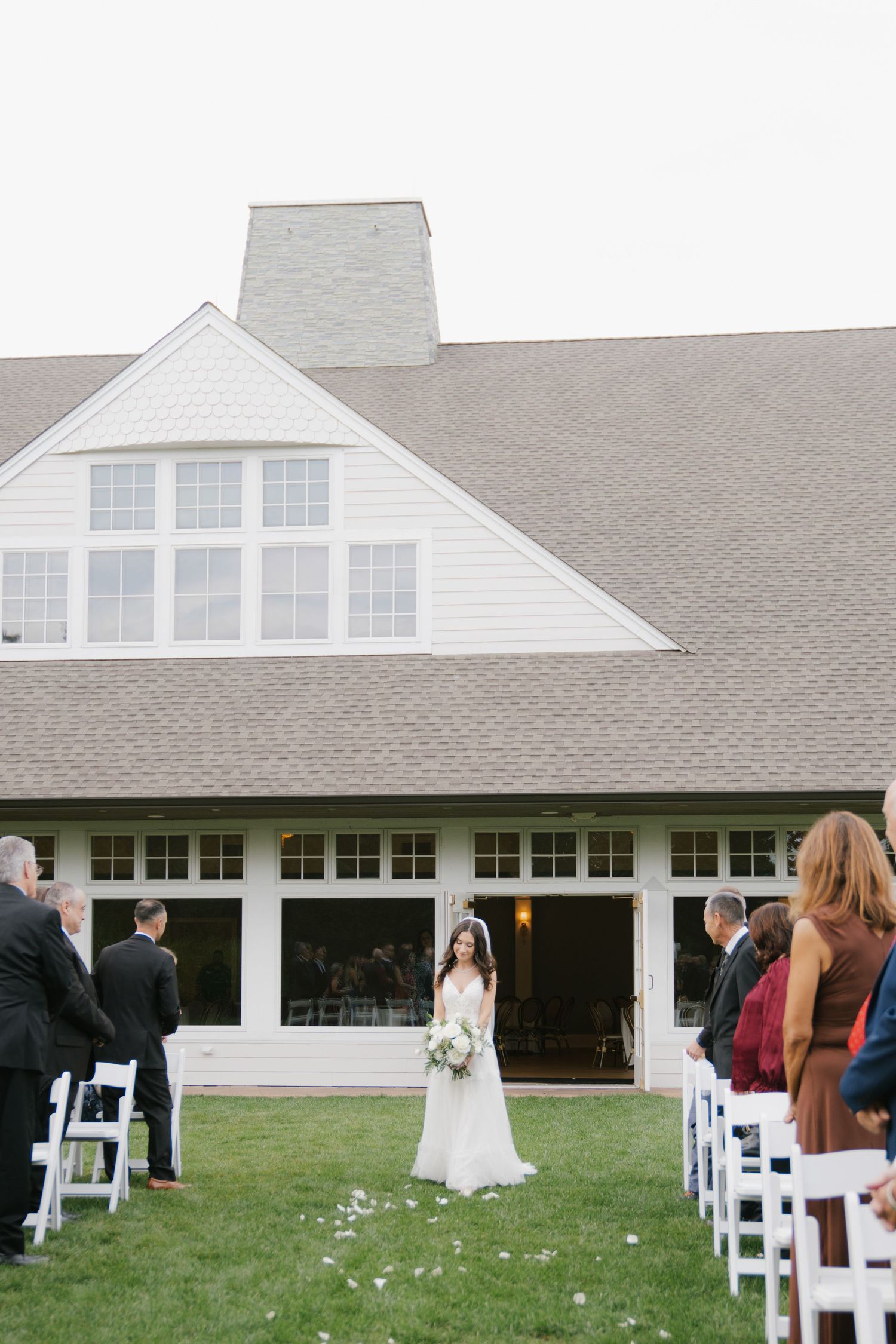 Elegant white wedding venue with gray roof features outdoor ceremony setup with white chairs and flower petals on grass lawn.