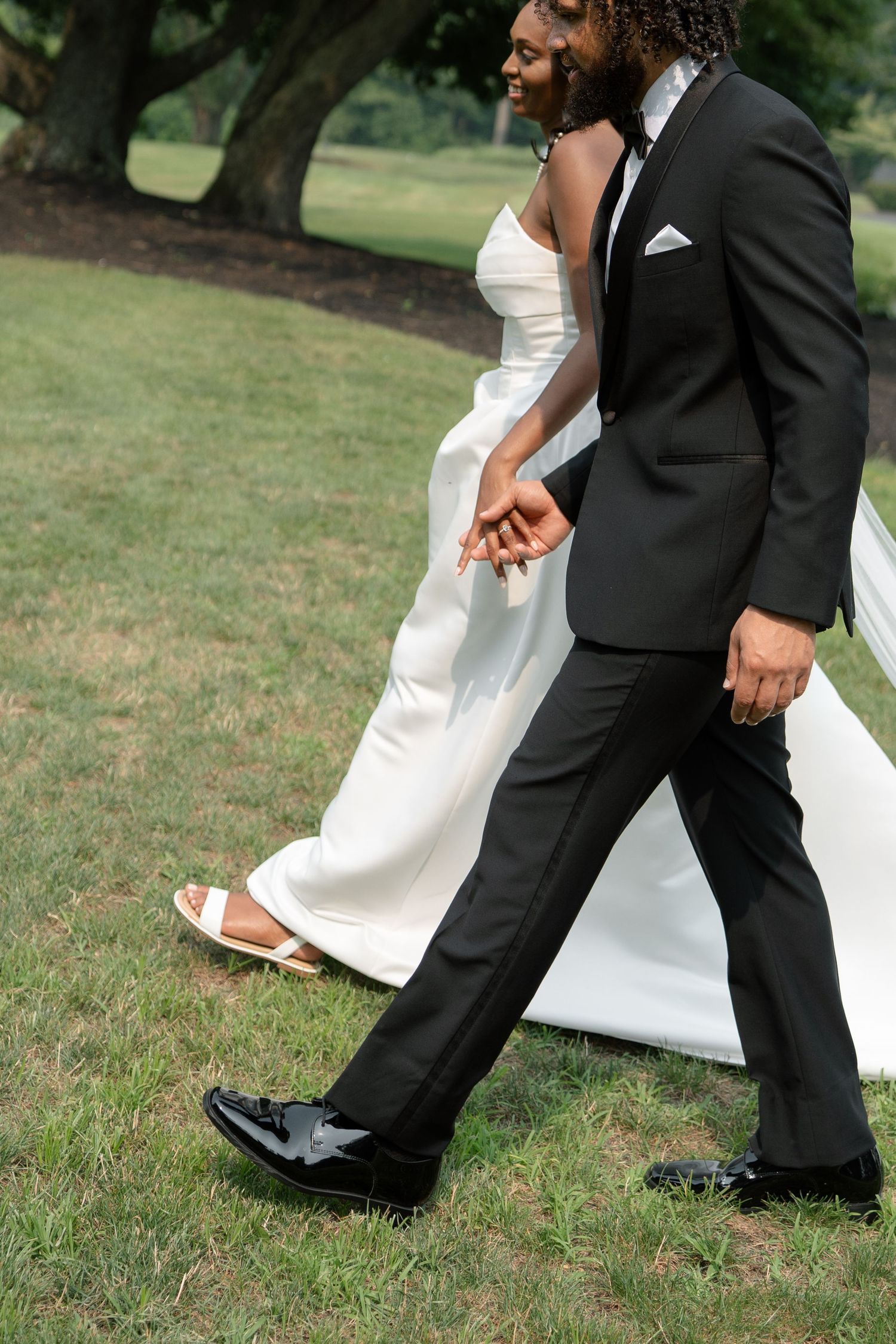 A Black bride and groom walk hand in hand on green grass, she in a white gown, he in a black tuxedo.