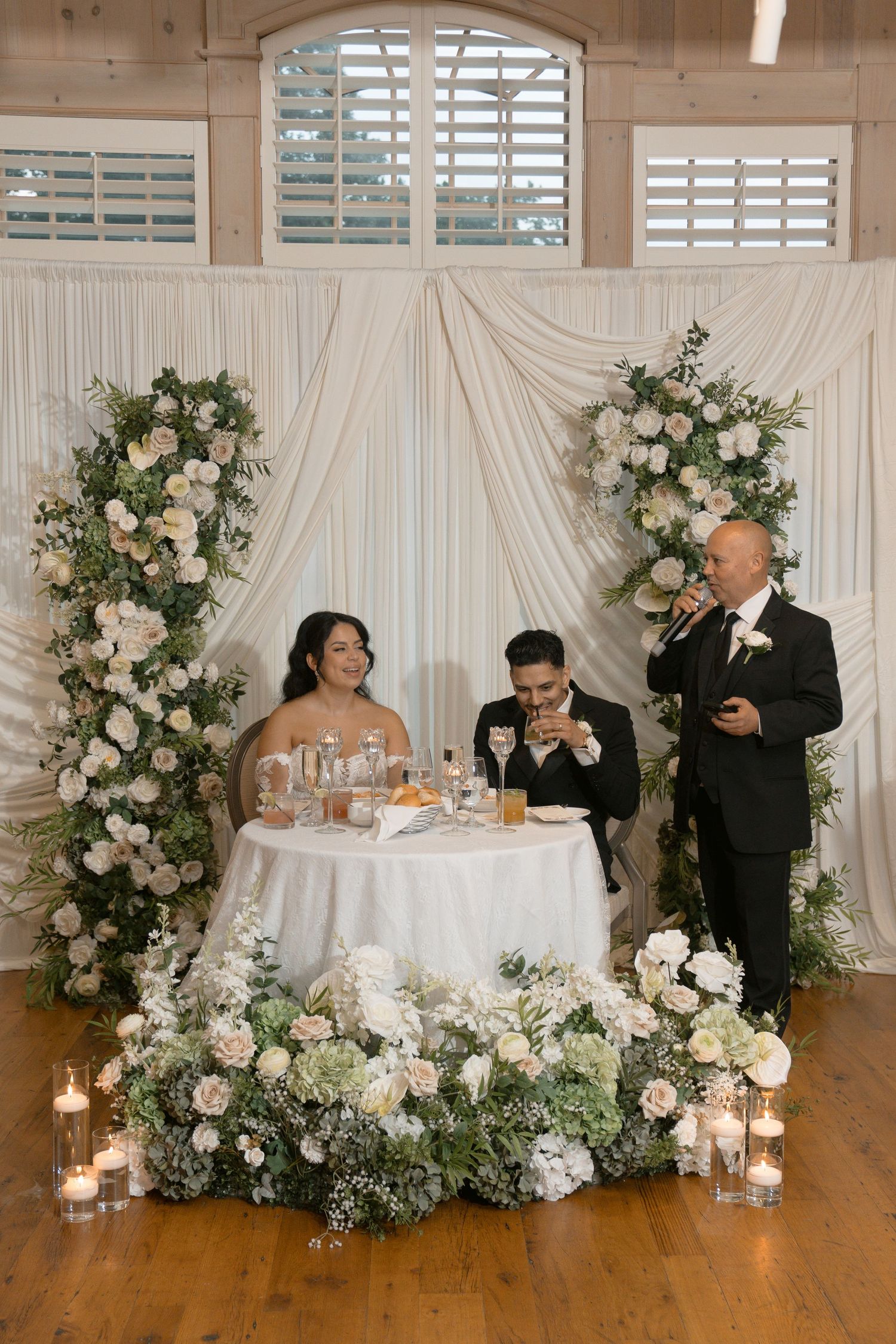 Bride and groom seated at sweetheart table with white floral arrangements and candles during wedding reception toast.