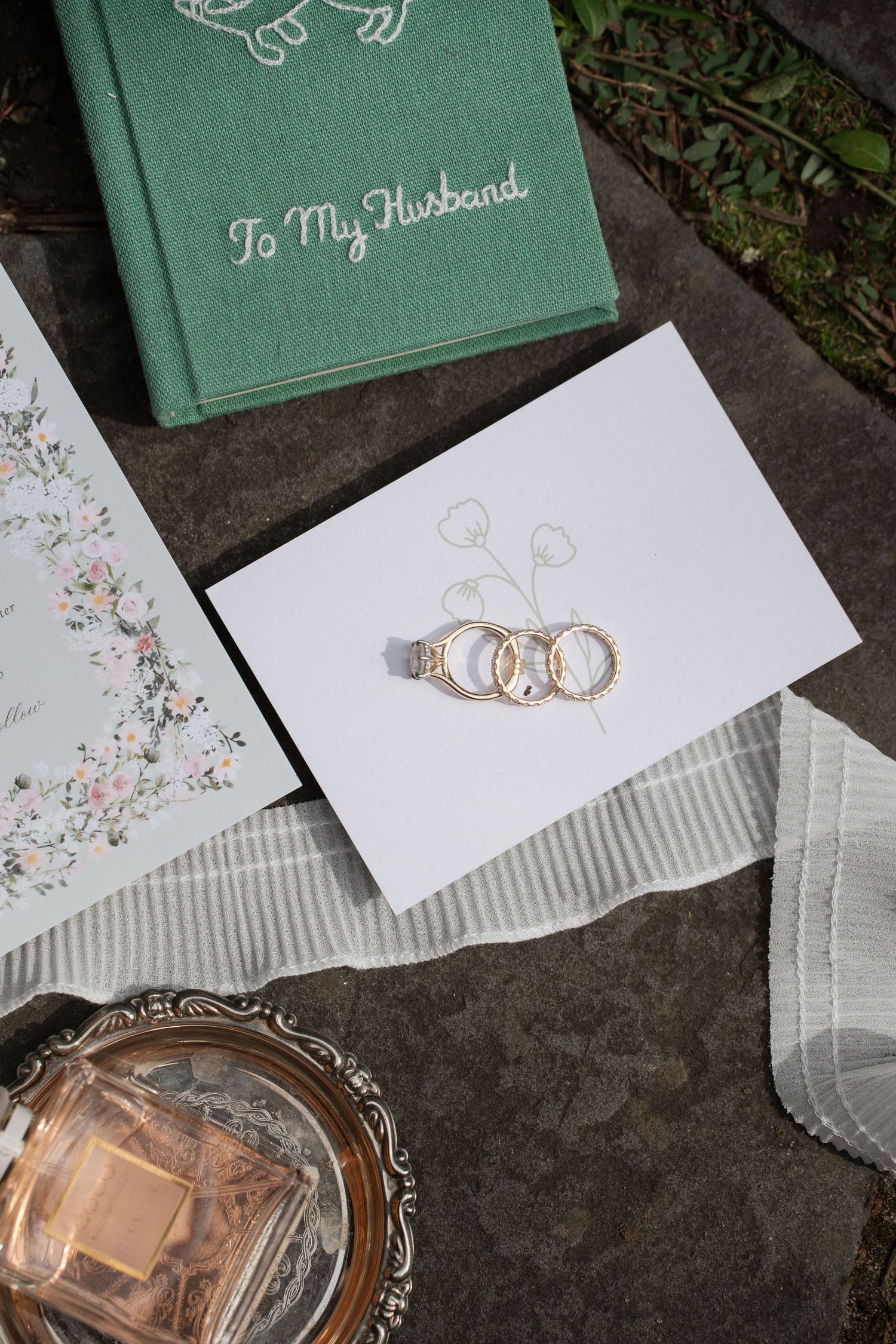 Wedding rings resting on a white card beside a green husband journal, floral stationery, perfume bottle, and sheer ribbon.