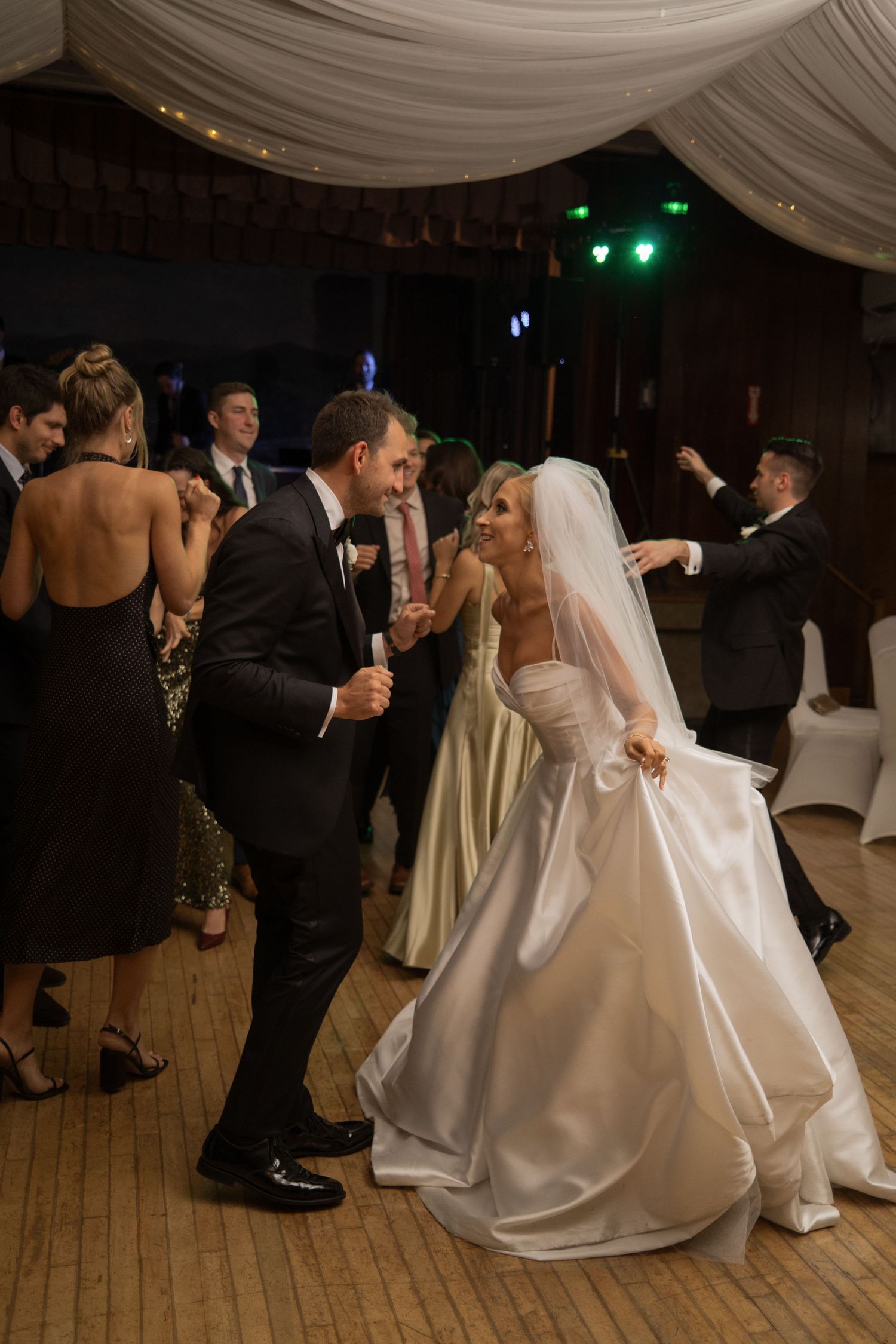 Bride and groom sharing romantic dance on crowded wedding reception dance floor under warm ambient lighting.