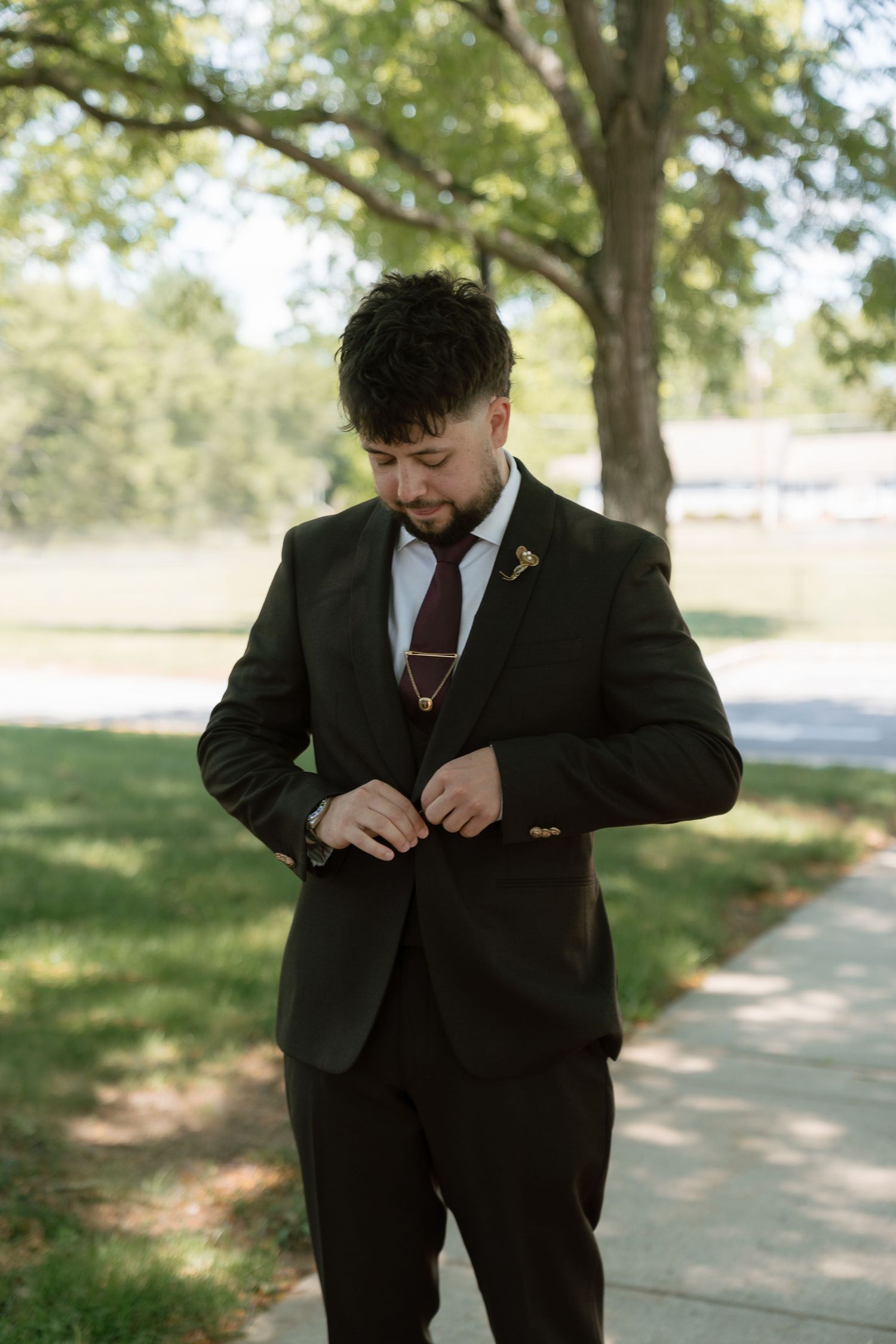 Groom in dark brown three-piece suit adjusting jacket button outdoors with green trees in background.