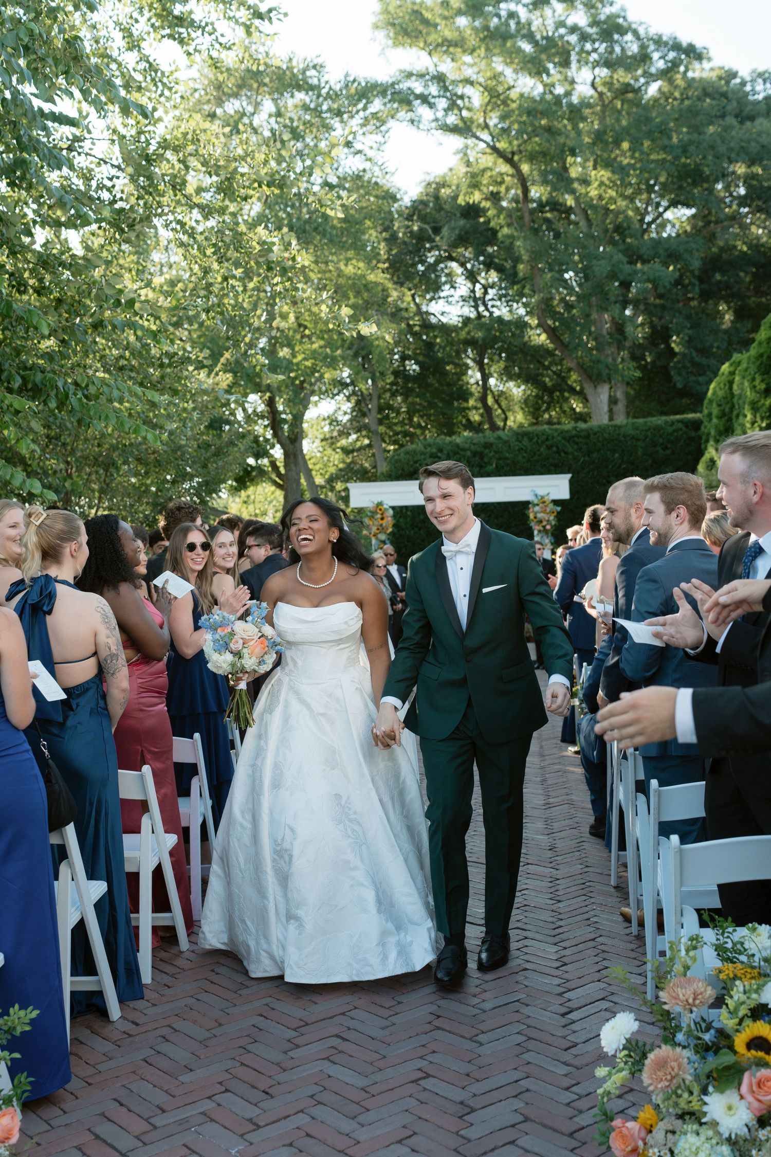 Joyful interracial newlyweds walk back down the aisle after outdoor wedding ceremony with guests.