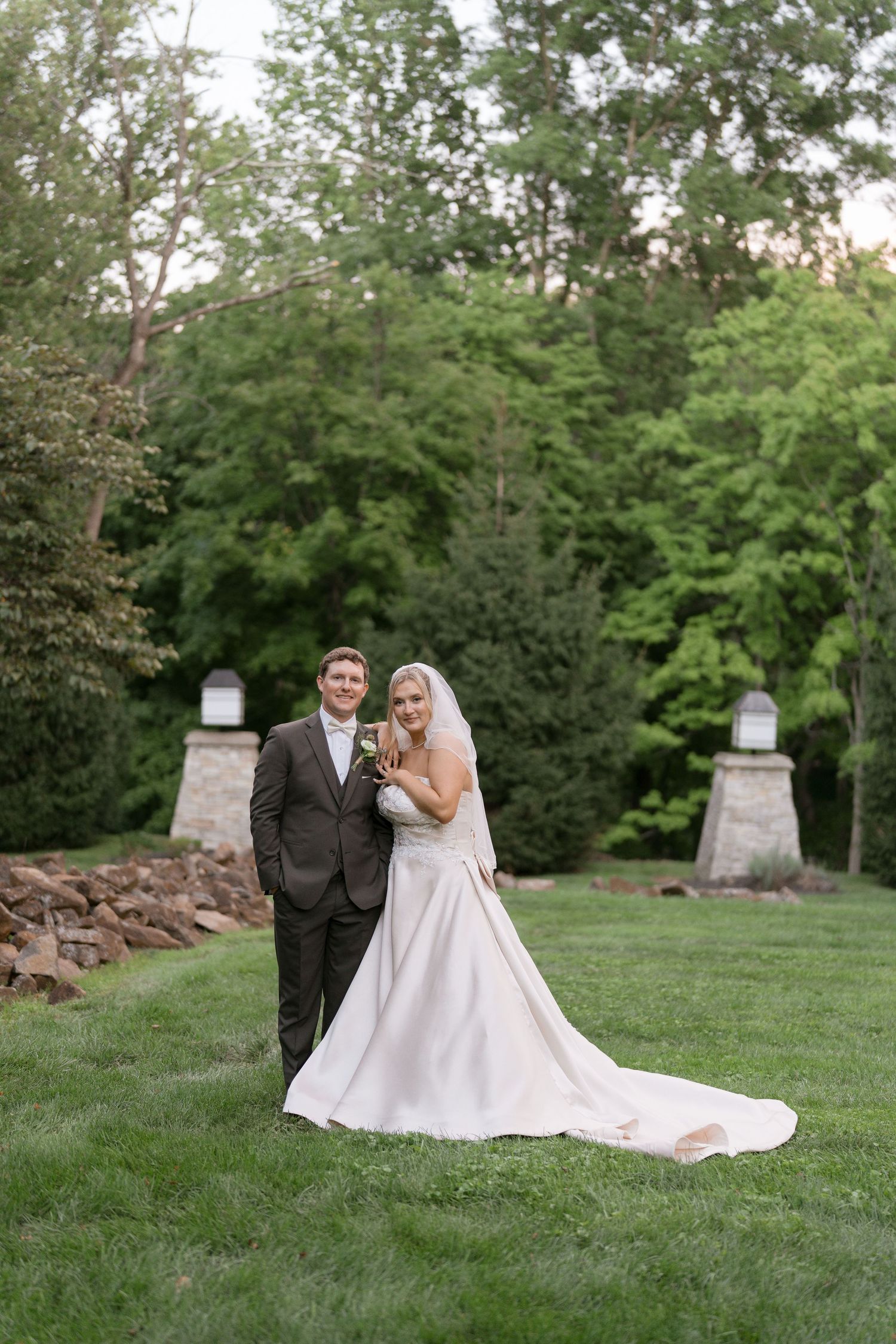 A bride and groom pose together on a lush green lawn surrounded by trees during their outdoor wedding ceremony.