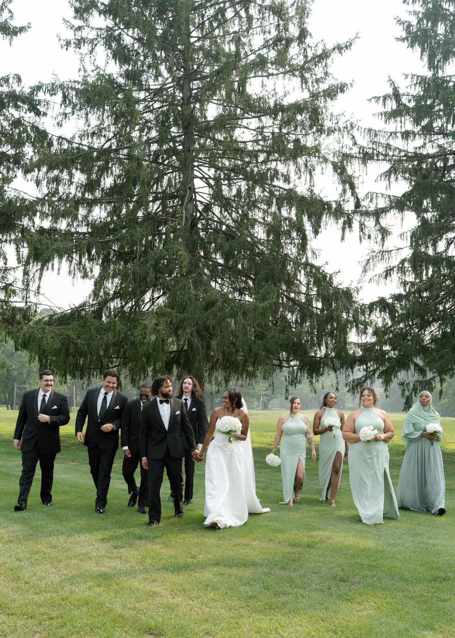 Wedding party walks across green lawn with large evergreen tree, bride in white gown with groomsmen and bridesmaids in sage.