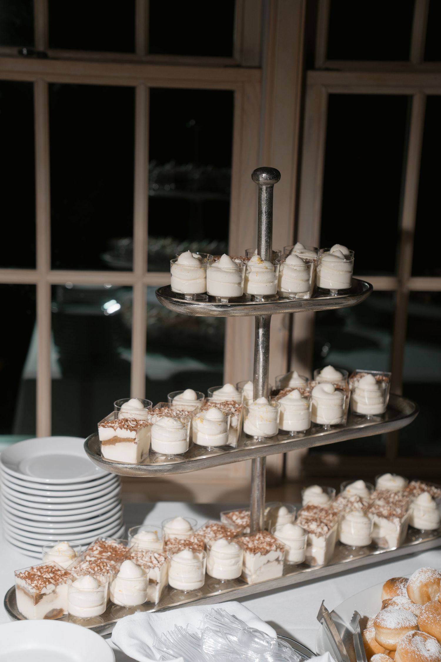 Three-tiered silver dessert stand displaying elegant white cream mini cakes and tiramisu bites at a wedding reception.