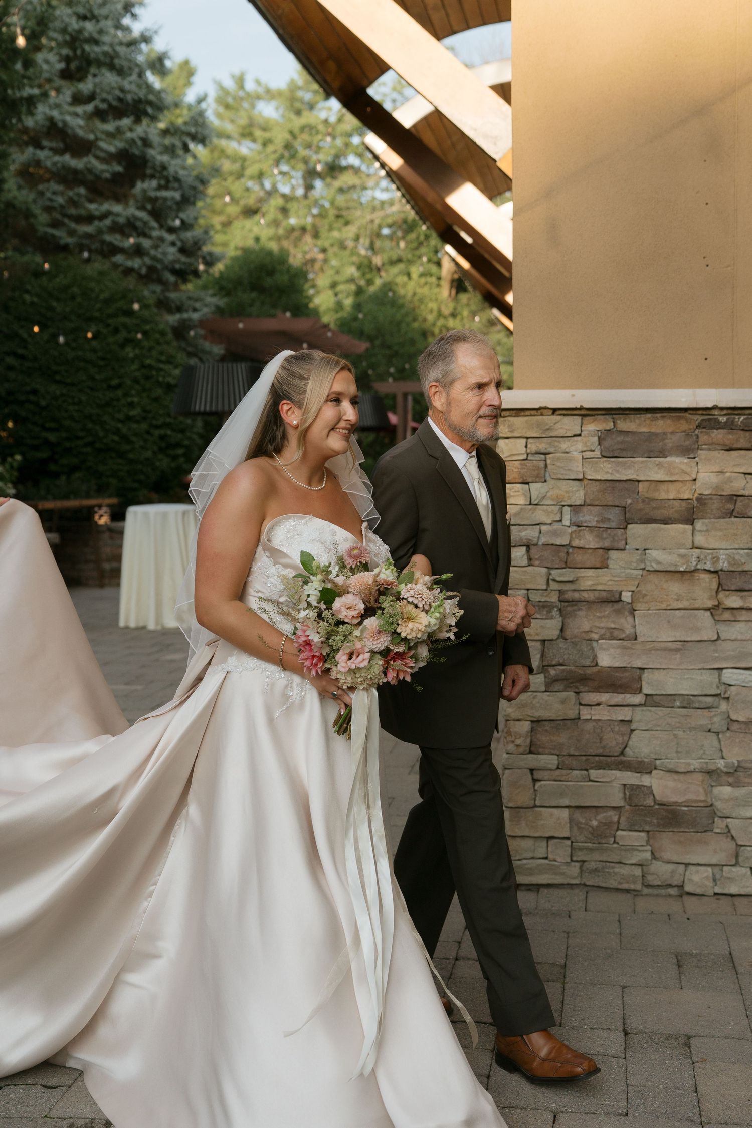 Smiling bride in a strapless satin gown holding a pink bouquet walks with a man at an outdoor wedding venue.