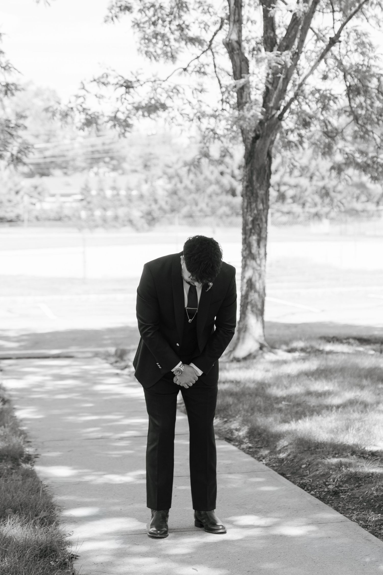 A groom in a black suit stands with bowed head on a garden path, hands clasped, in a black and white photo.