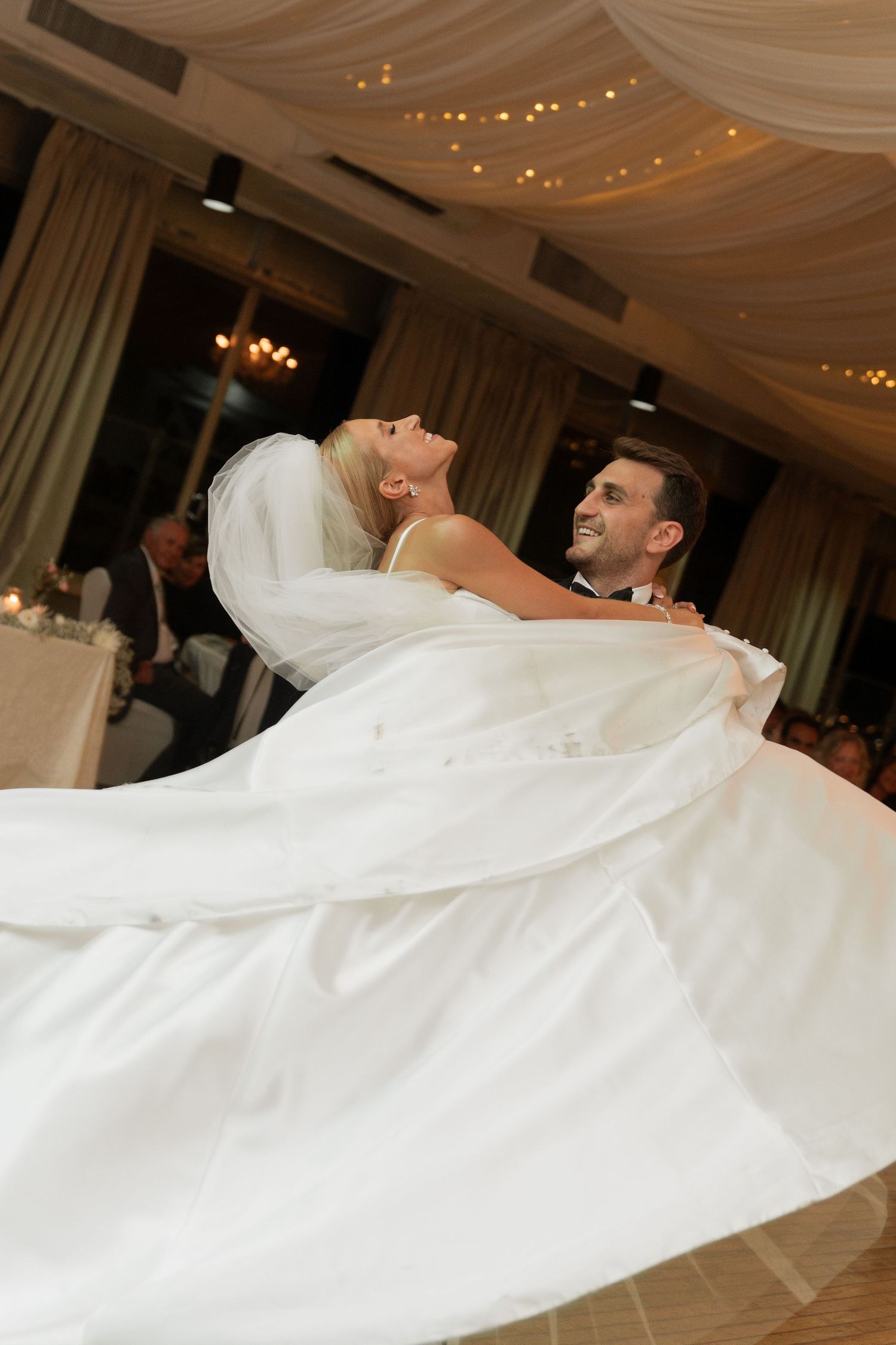 Groom lifts bride in white ballgown during joyful first dance at softly lit wedding reception.
