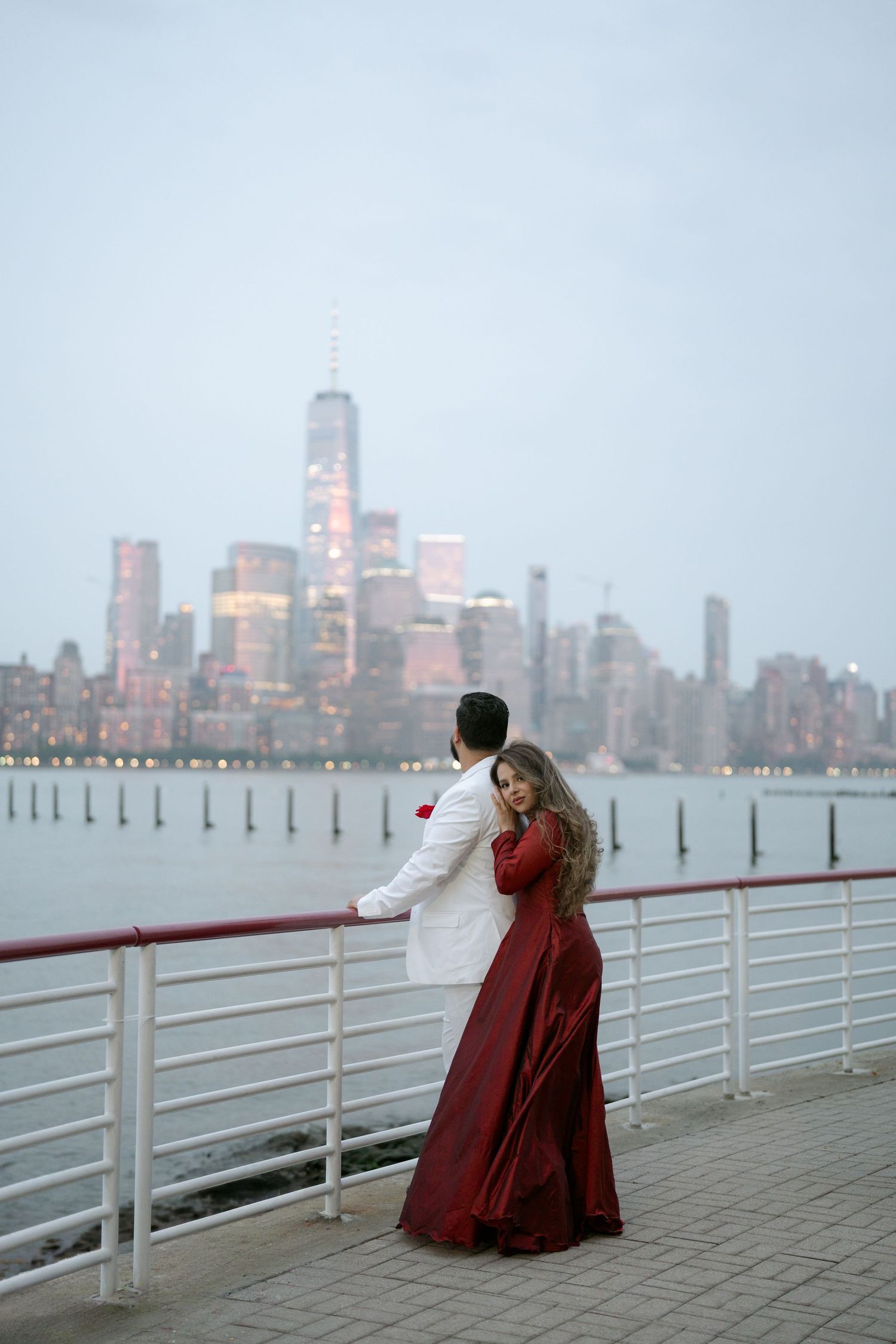 Couple in elegant red gown and white suit embracing on waterfront pier with NYC skyline at dusk.